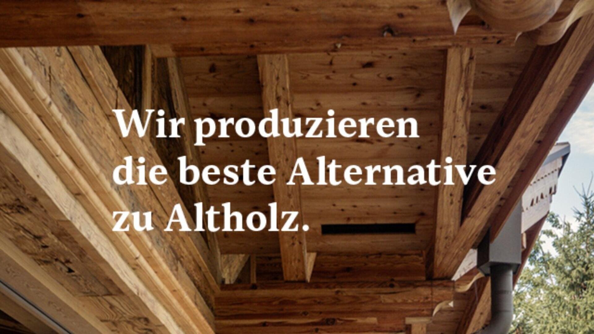 Wooden ceiling with exposed beams and carved details. Overlaid German text reads: Wir produzieren die beste Alternative zu Altholz. (English: We produce the best alternative to old wood.).