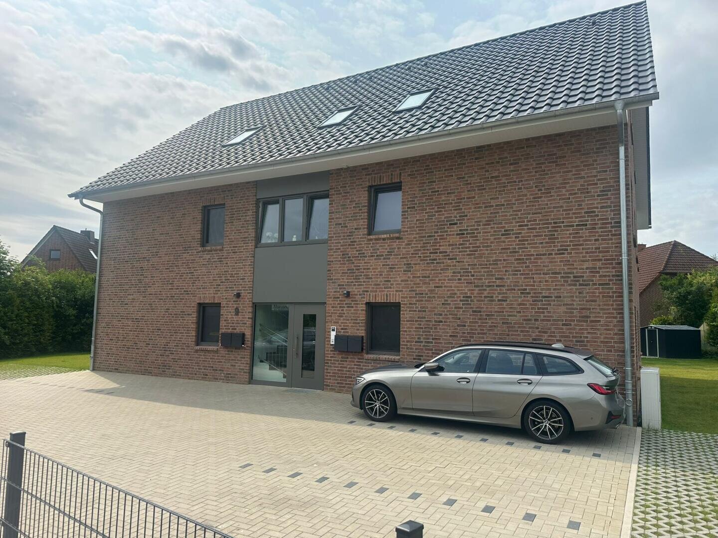 Ein modernes, zweistöckiges Backsteinhaus mit Schrägdach und Oberlichtern. Ein silberner Kombi ist in der gepflasterten Einfahrt vor dem Haus geparkt, im Hintergrund sind ein Rasen und die Nachbarhäuser zu sehen.