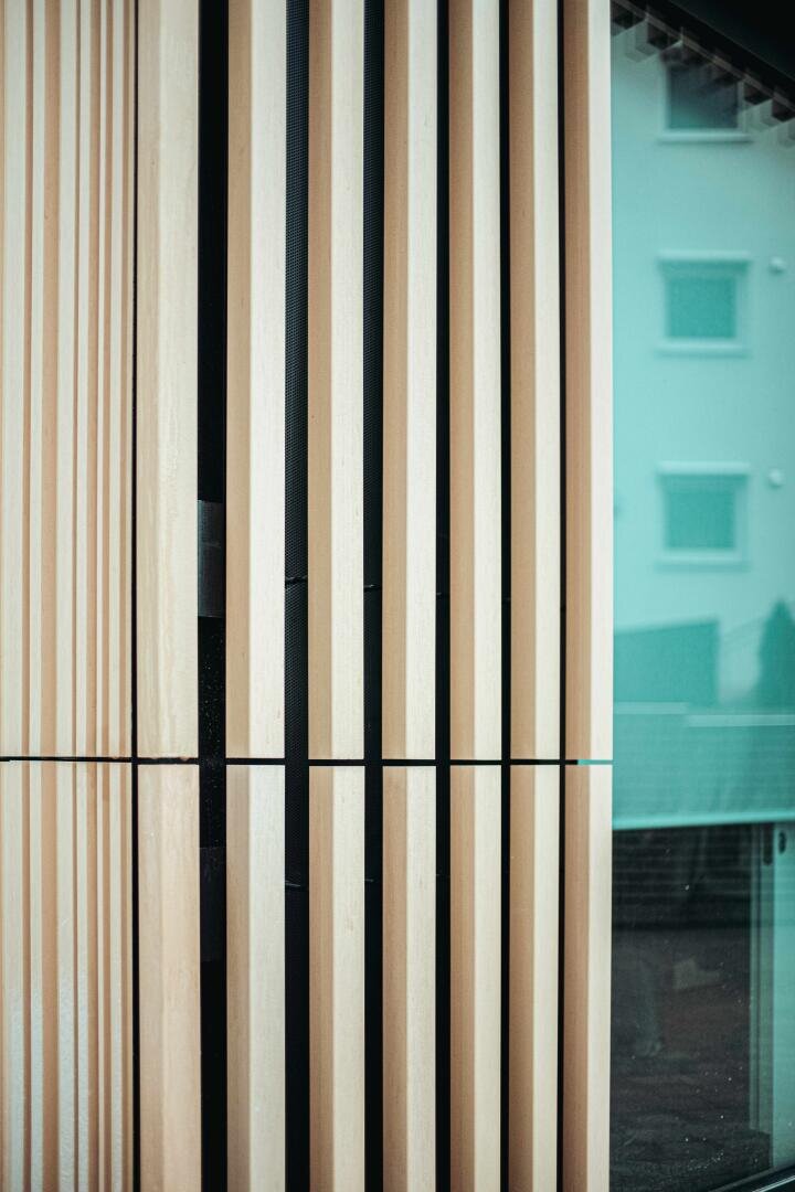 Close-up of vertical wooden slats forming part of a modern architectural façade, with a building reflected in a glass surface on the right.