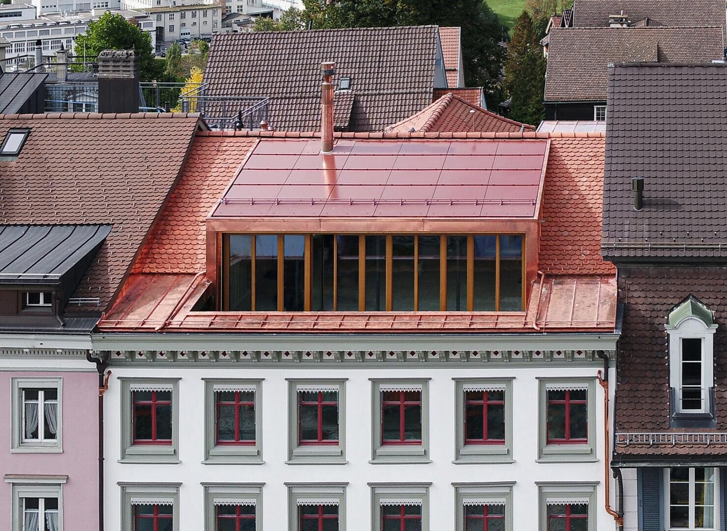 A renovated building with a red tiled roof and large modern skylight windows, surrounded by traditional buildings in an urban neighborhood. The façade features green trim and red-framed windows.