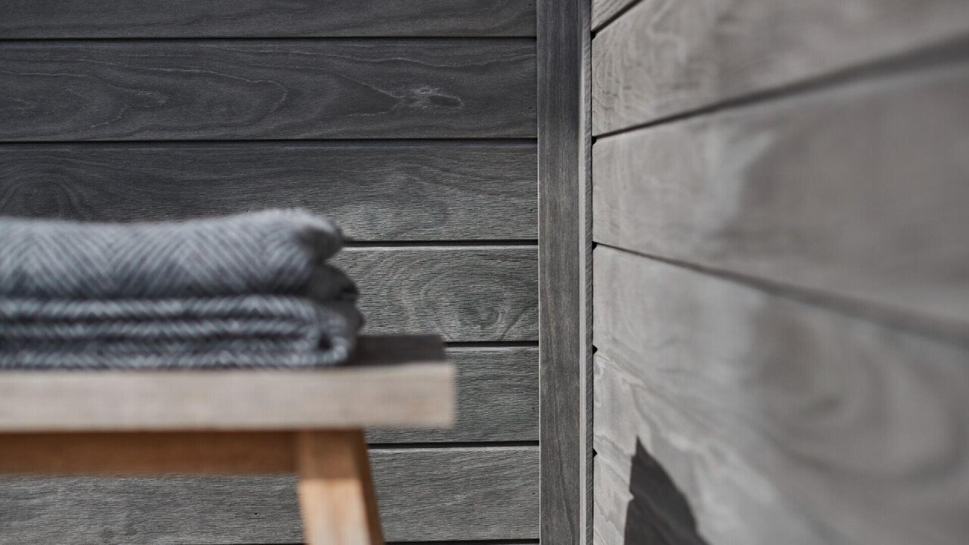 A neatly folded gray blanket sits on a wooden bench against a backdrop of smooth, gray wooden wall panels with a subtle grain pattern, creating a modern and minimalistic look.