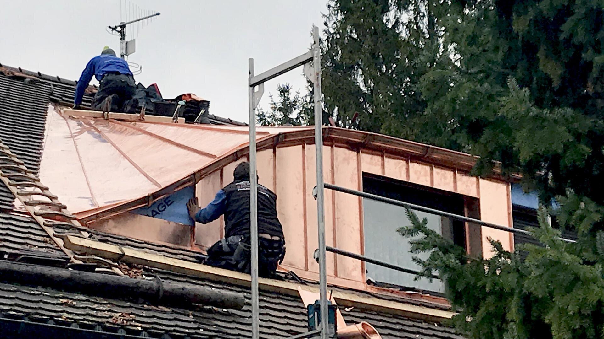 Zwei Arbeiter montieren Kupferplatten auf einem Dachfenster. Einer kniet auf dem Dach, der andere arbeitet am Fenster. In der Nähe sind Gerüste und Bäume zu sehen.