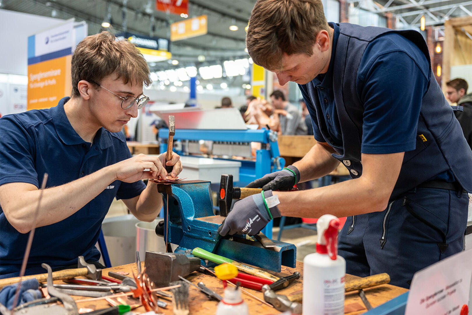 Zwei junge Männer in Arbeitsuniformen arbeiten mit Handwerkzeugen an Metallteilen an einem Werkstatttisch und konzentrieren sich dabei auf die Arbeit. Um sie herum sind verschiedene Werkzeuge und Ausrüstungen in einer belebten Halle zu sehen.