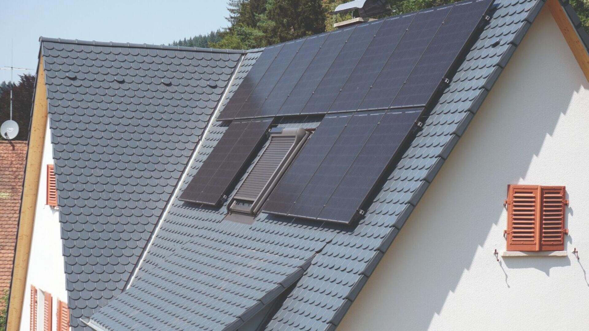 A house with dark gray roof tiles has solar panels installed on the sloped roof. The house features red window shutters and is surrounded by trees and greenery.