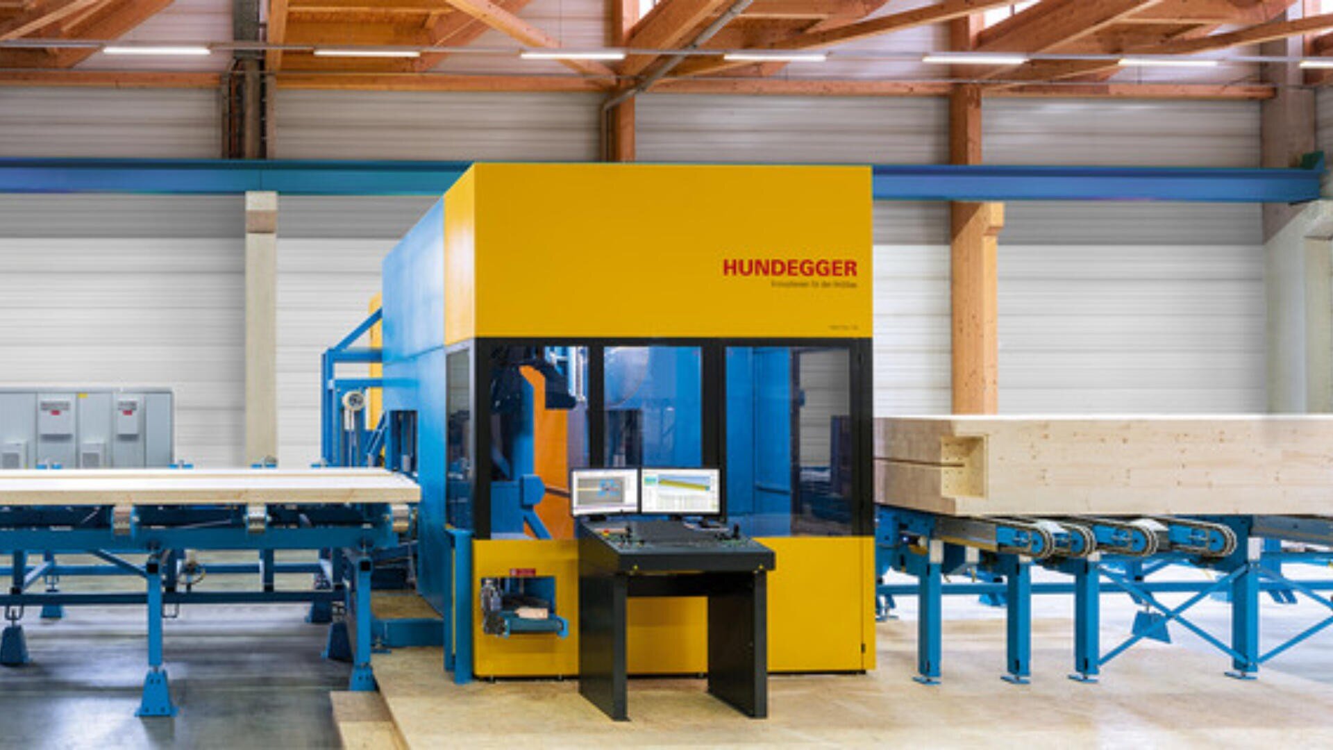 Eine große gelb-blaue Hundegger-Holzbearbeitungsmaschine steht in einer Fabrik mit Holzbalken auf Förderbändern und einem Computerarbeitsplatz davor. Die Fabrik hat Holzbalken an der Decke und weiße Wände.