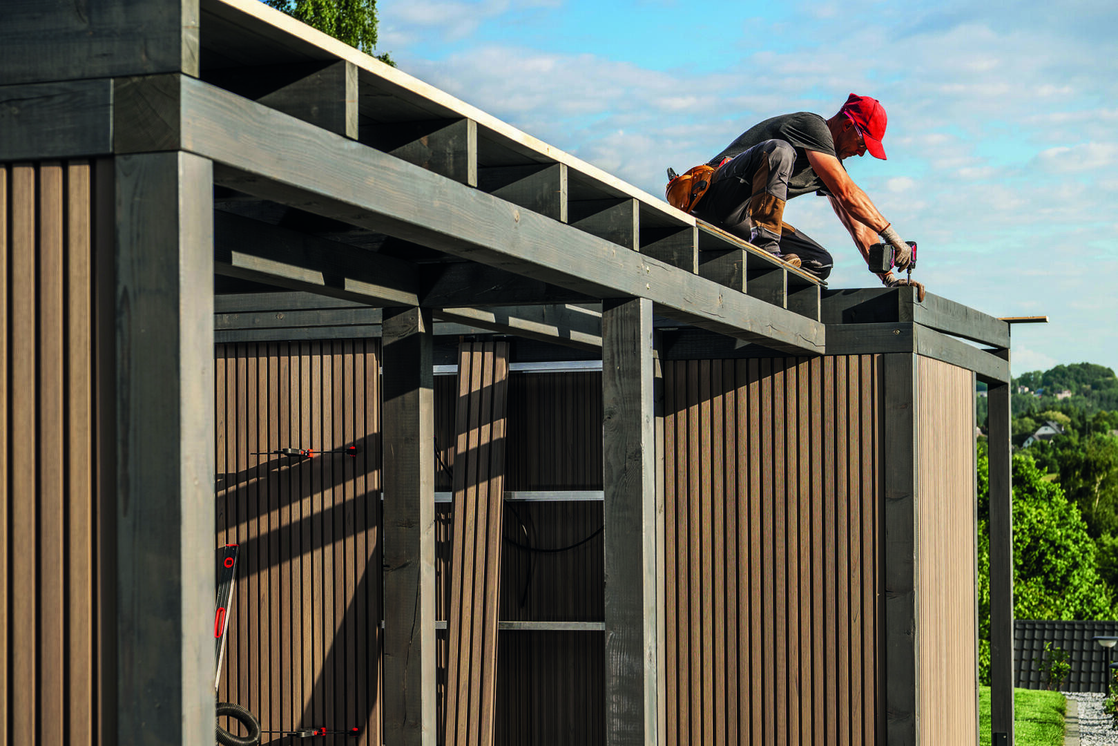 A person wearing a red cap kneels on the roof of a partially constructed wooden shed and uses a power tool. The shed is outdoors on a sunny day, surrounded by greenery.