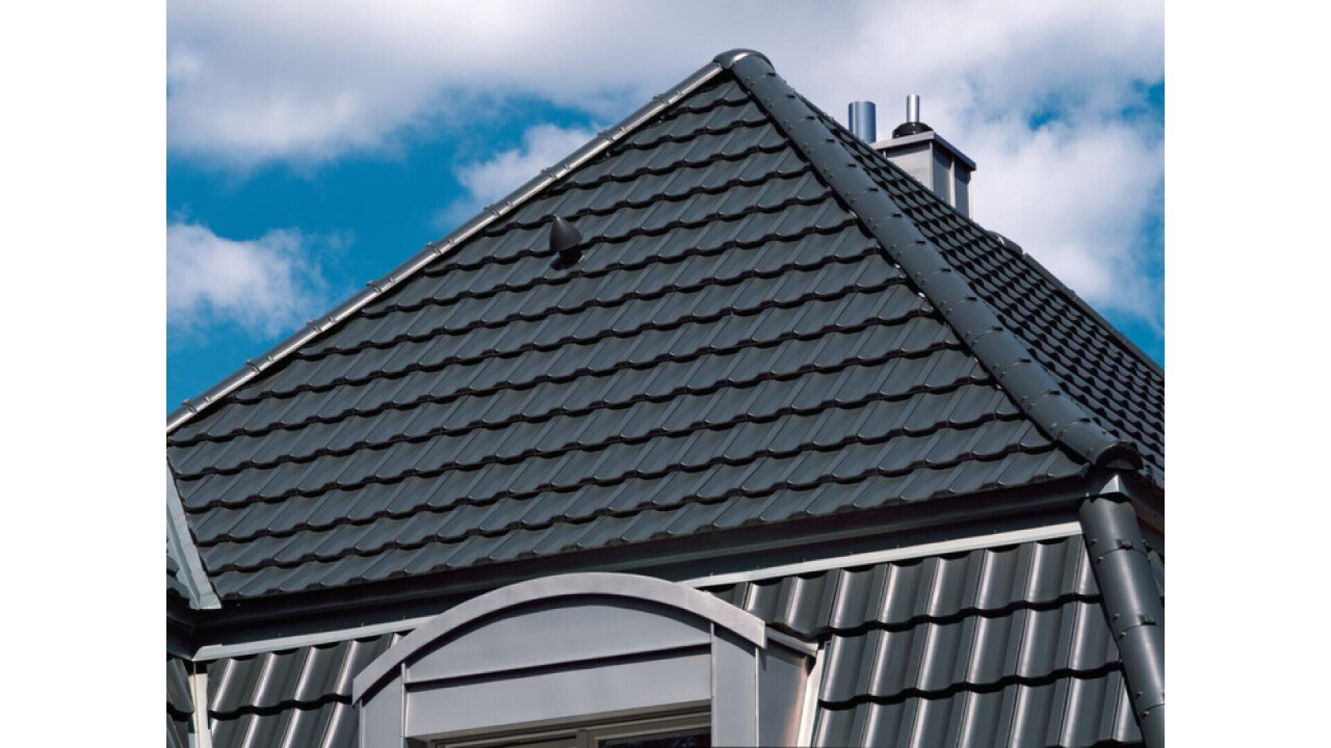 A close-up view of a house roof with dark gray, metal tile shingles, featuring a dormer window and a chimney against a blue sky with scattered white clouds.