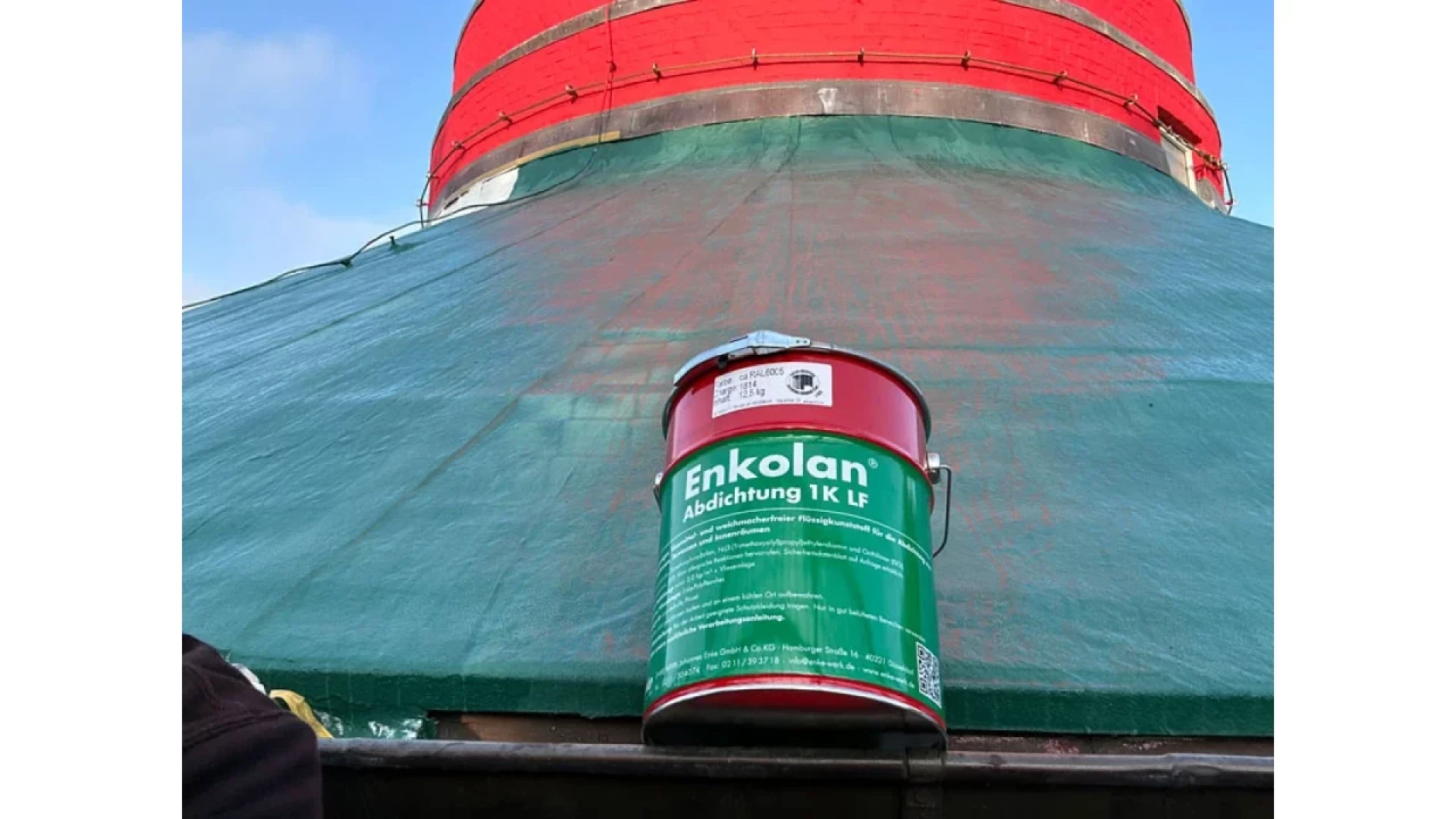 A large green and red Enkolan waterproofing bucket sits on a ledge in front of a conical structure covered with green netting and topped with a red section under a blue sky.
