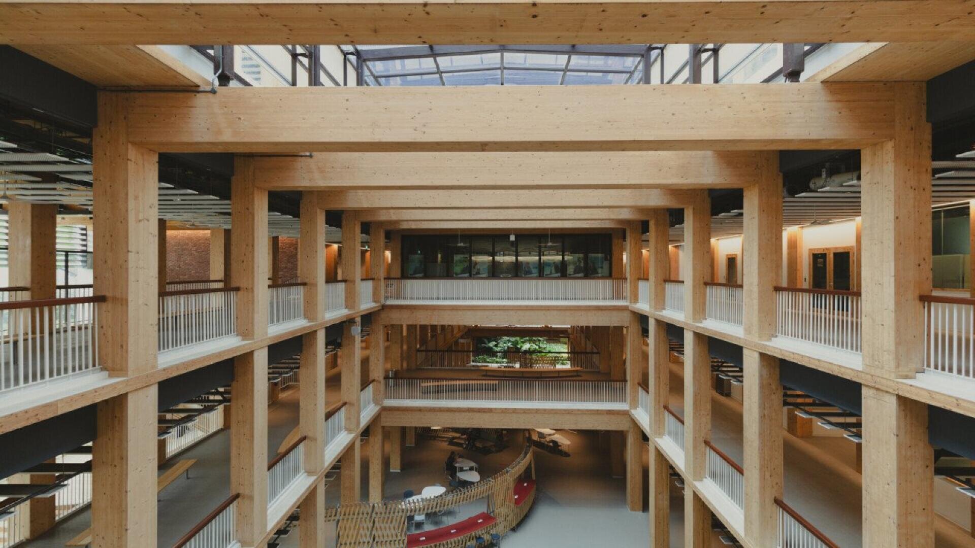 Interior view of a modern, multi-story building with exposed wooden beams, open walkways, glass walls, and natural light streaming in from a skylight above. The space features minimalist design and indoor plants.