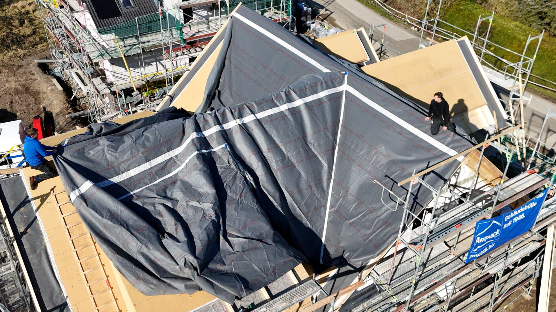 Luftaufnahme eines im Bau befindlichen Gebäudes mit Baugerüst. Das Dach ist mit einer großen grauen Plane abgedeckt, und zwei Personen stehen auf beiden Seiten der Struktur. Baumaterialien und Werkzeuge sind sichtbar.