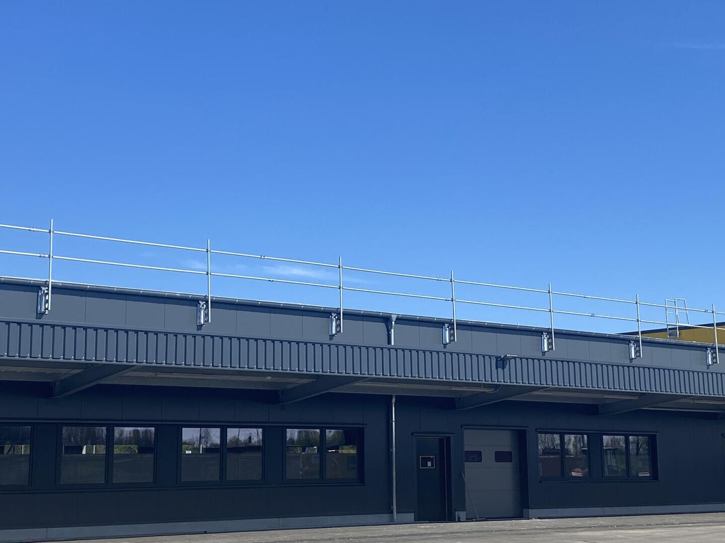 Ein Industriegebäude mit Flachdach, dunkelgrauen Wänden und mehreren Fenstern, mit einem Sicherheitsgeländer entlang der Dachkante, unter einem klaren blauen Himmel.
