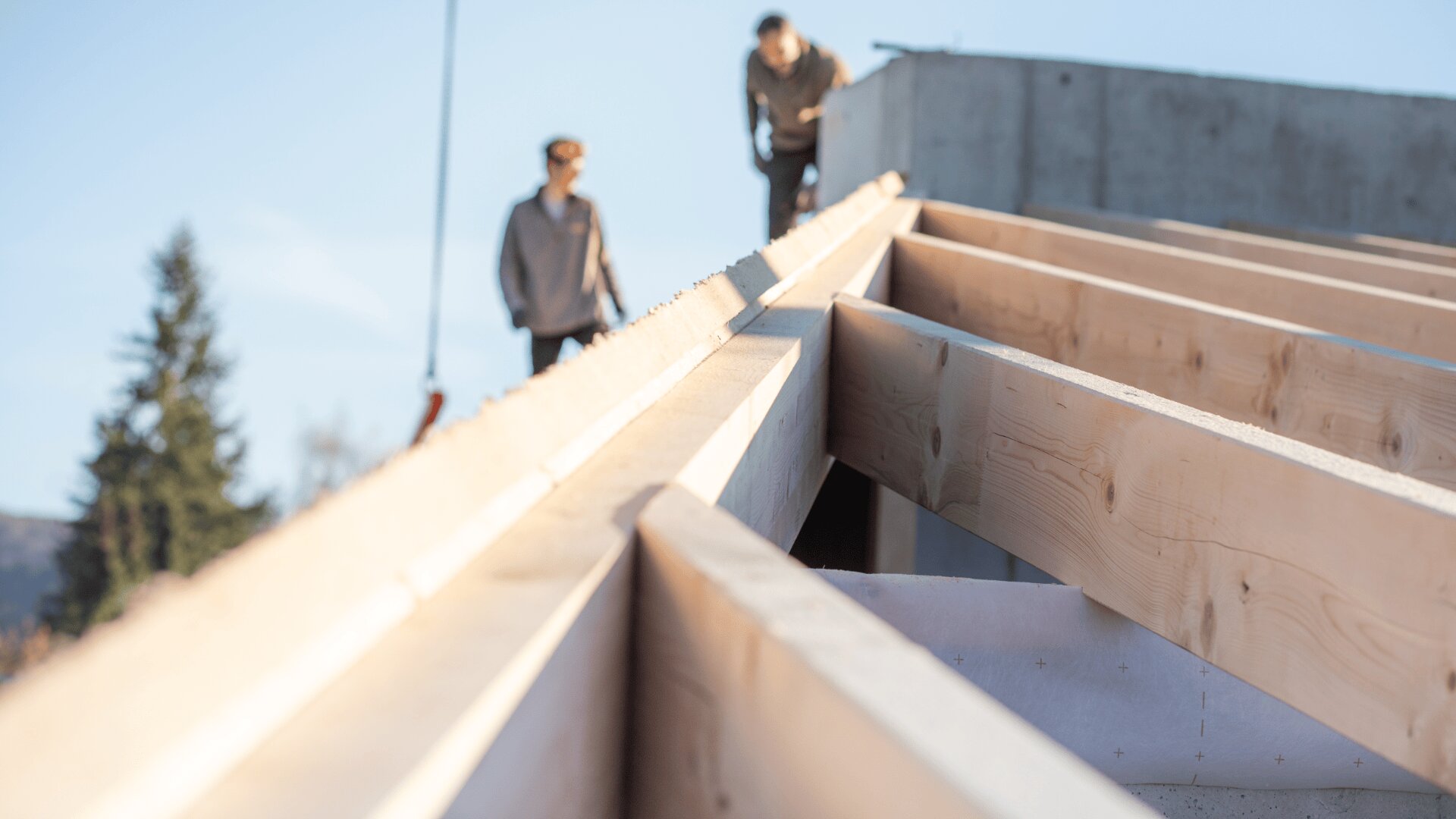 Close-up of wooden roof beams under construction with two blurred people in the background. Clear sky and a tree are visible, suggesting an outdoor building site.