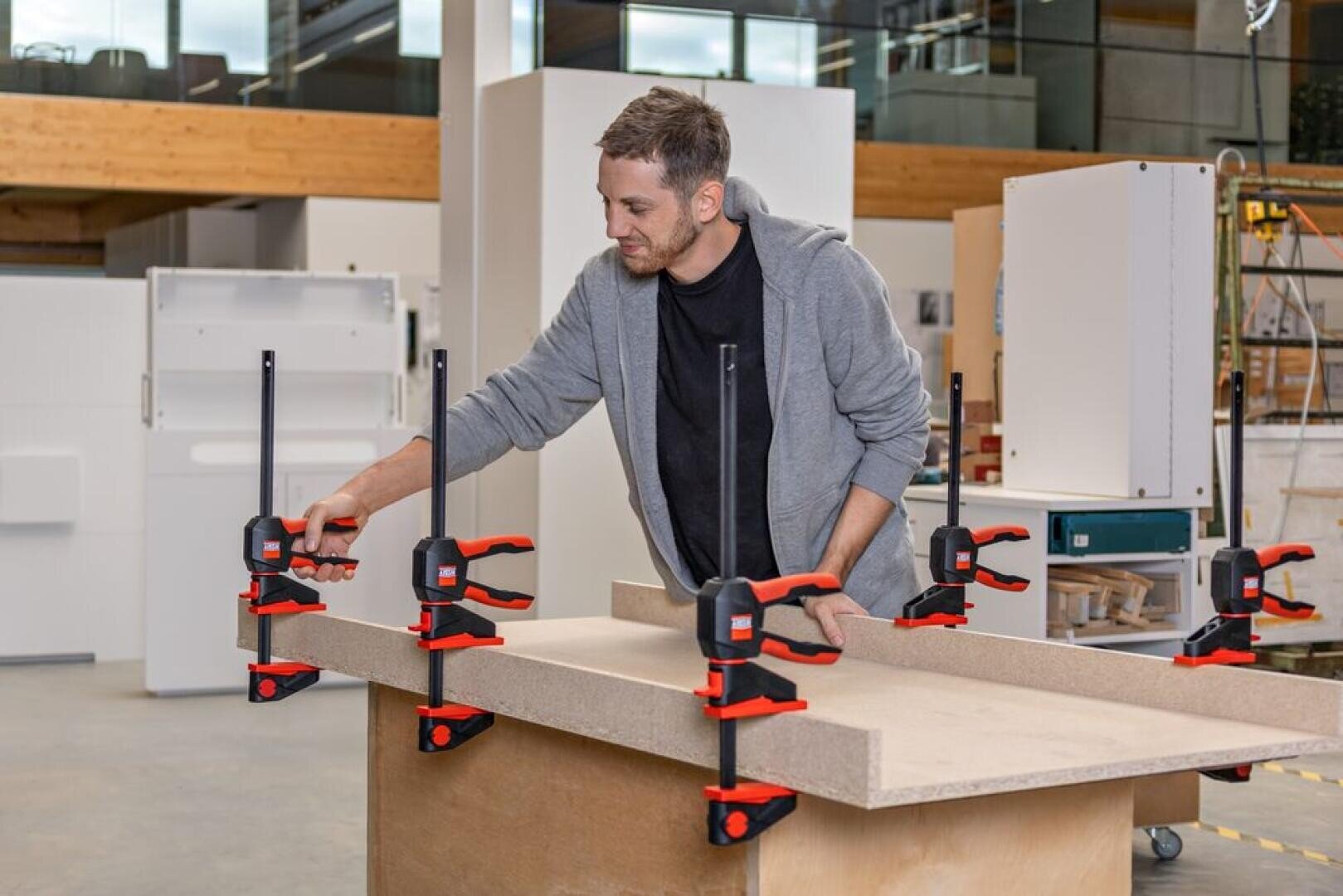 Ein Mann in einer Werkstatt benutzt mehrere rote und schwarze Zwingen, um zwei große Holzplatten auf einer Werkbank zu befestigen, die von Schränken und Werkzeugen umgeben ist.