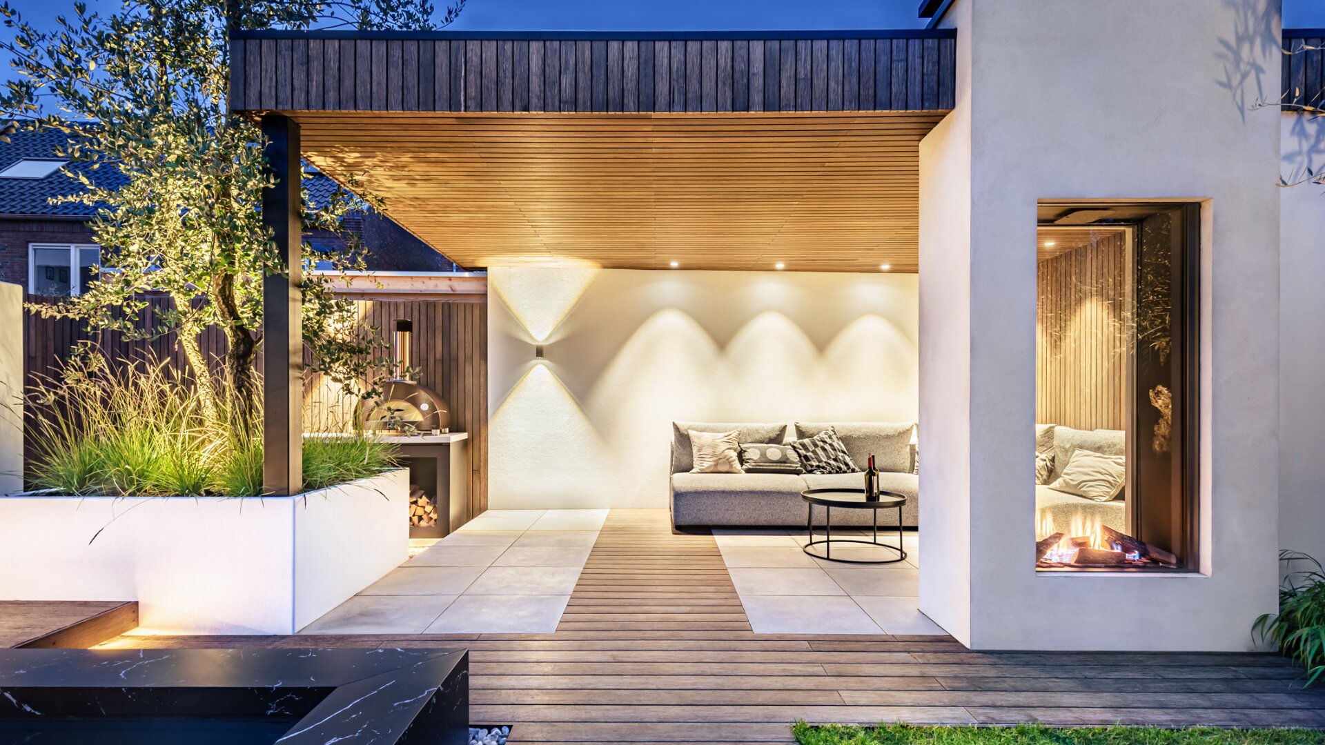 Moderne, überdachte Terrasse mit Holzdecke, Einbauleuchten, einem Gartensofa, einem kleinen runden Tisch, einem eingebauten Kamin und üppiger Gartengestaltung. Am frühen Abend ist der Raum elegant beleuchtet.