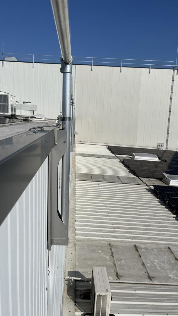 Blick von einem Dach mit Metallverkleidung, einem Metallrohr, das am Rand entlangläuft, Sonnenkollektoren, Dachmaterial und Klimaanlagen unter einem klaren blauen Himmel.