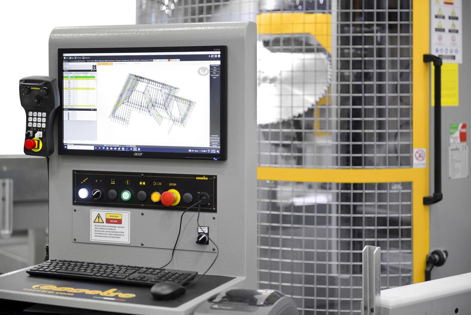 Eine Nahaufnahme des Bedienfelds einer Industriemaschine mit einem Monitor, auf dem ein 3D-Diagramm, eine Tastatur, Not-Aus-Tasten und im Hintergrund ein Sicherheitskäfig angezeigt werden, der die Maschine umschließt.