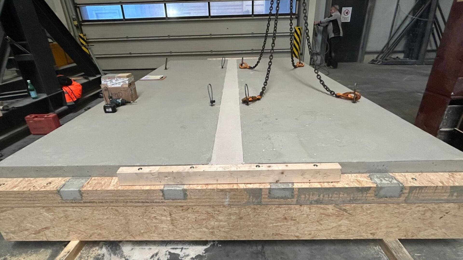 Große Betonplatte mit eingebetteten Metallstangen und Ketten zum Heben, die auf einer hölzernen Stützstruktur in einem Industrielager ruhen. Im Hintergrund steht eine Person in der Nähe der Ausrüstung.