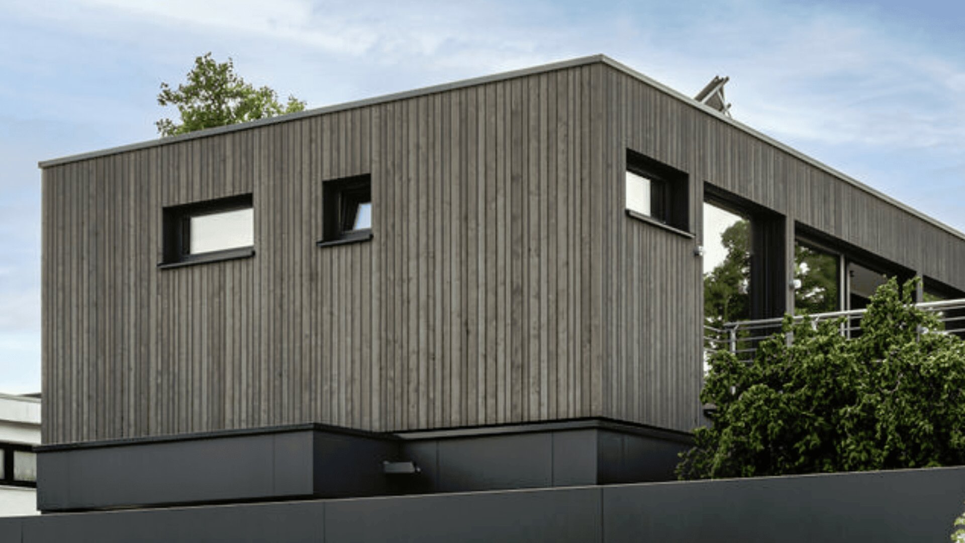 Ein modernes Haus mit vertikaler Holzverkleidung und dunkel gerahmten Fenstern, einem Flachdach und umgeben von Bäumen und Grün.