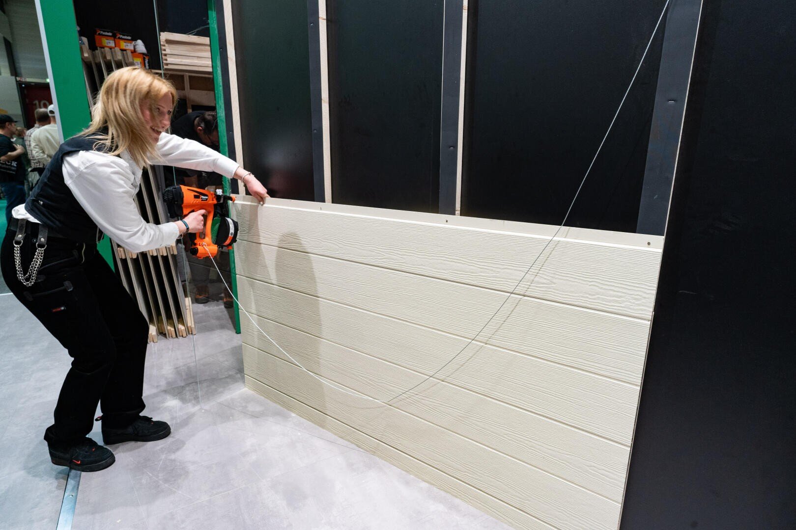 A person uses a nail gun to attach beige-colored wooden cladding panels to a black-framed wall structure, while tools and building materials can be seen in the background.