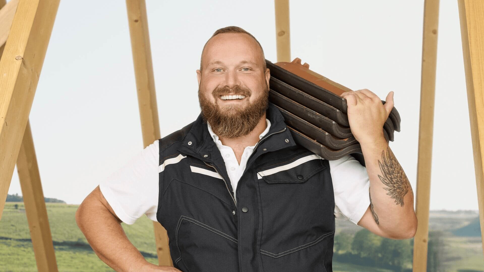 Ein lächelnder Mann mit Bart, einer schwarzen Weste über einem weißen Hemd, hält mehrere Dachziegel auf seiner Schulter. Er steht vor einer Holzrahmenkonstruktion, im Hintergrund sind grüne Felder zu sehen.