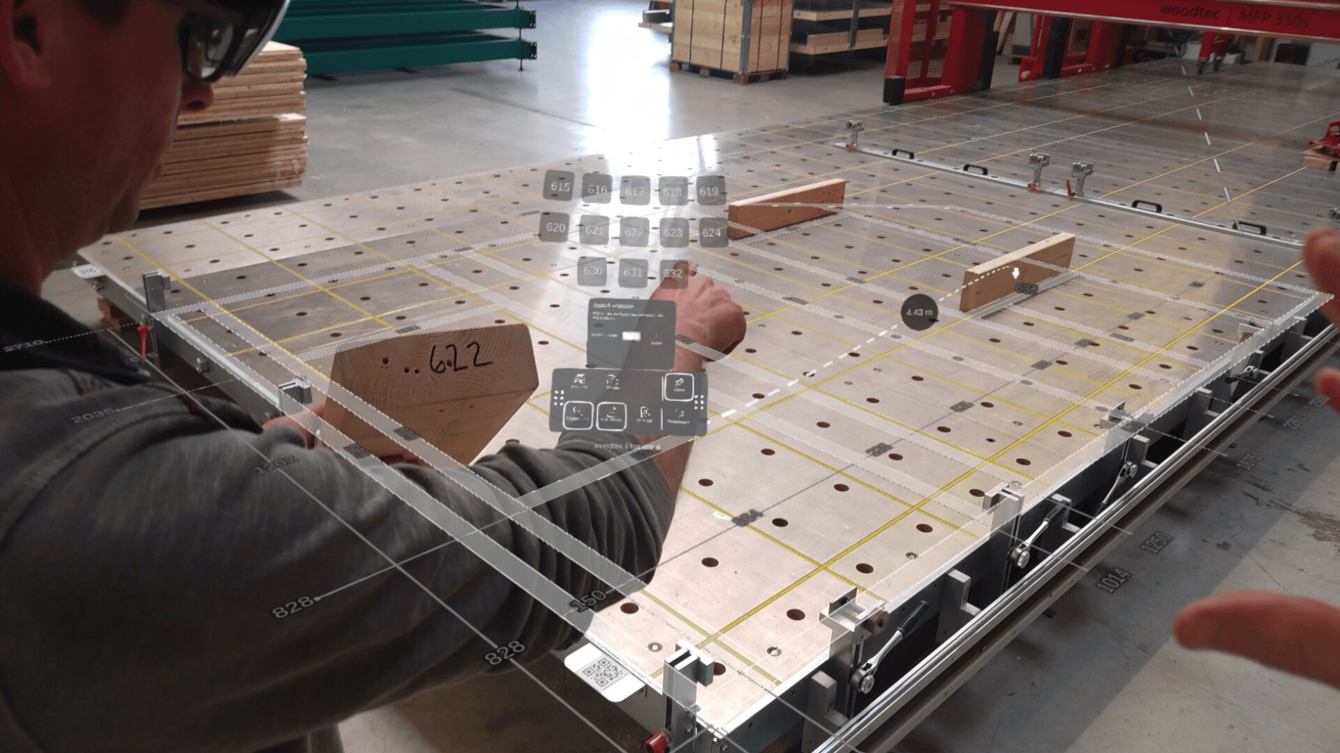 Eine Person verwendet eine Augmented-Reality-Brille, um digitale Bedienelemente und Diagramme anzuzeigen, die auf einer großen hölzernen Montagewerkbank in einer Fabrik oder Werkstatt eingeblendet sind.