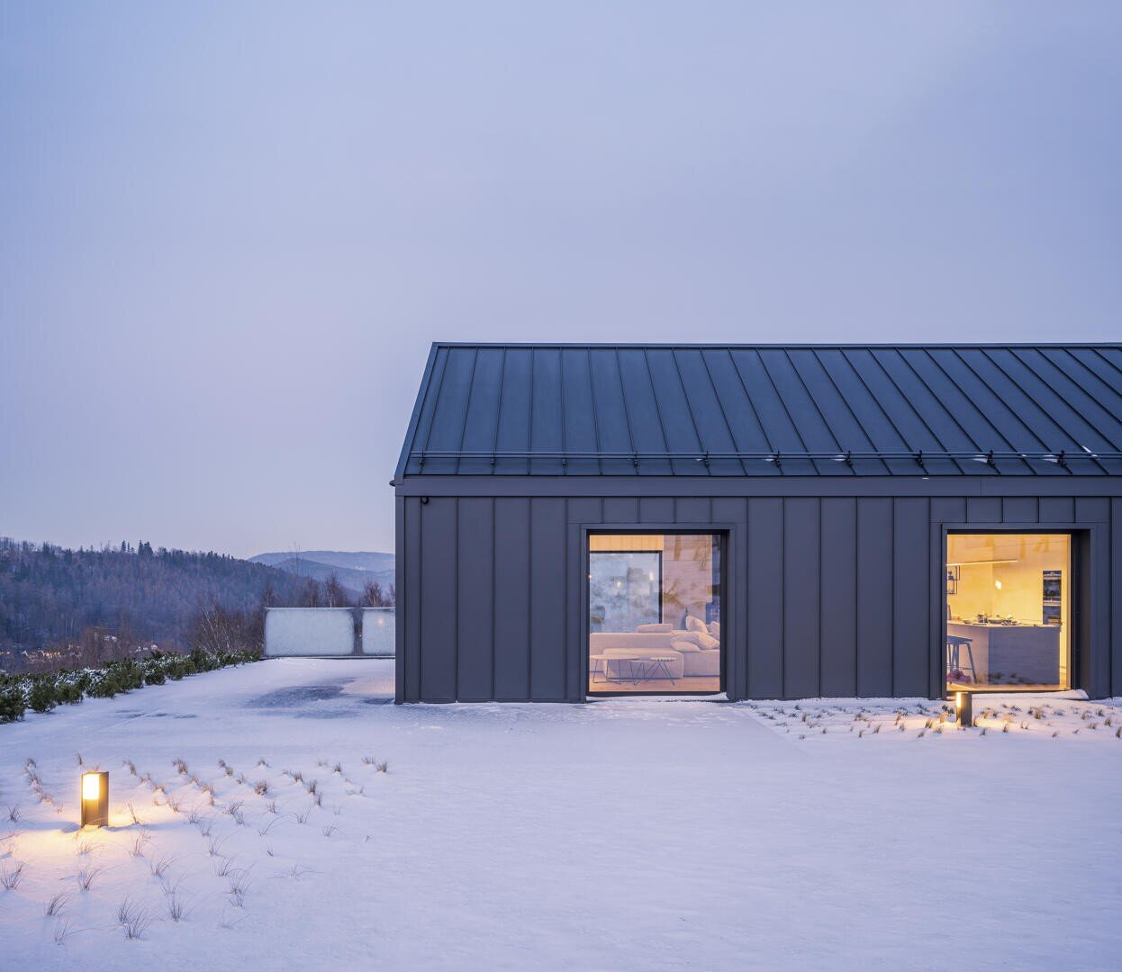 A modern house with a dark metal roof and two large, illuminated windows stands in a snow-covered courtyard at dusk. The outdoor lighting gently illuminates the snow, and trees can be seen in the distance.