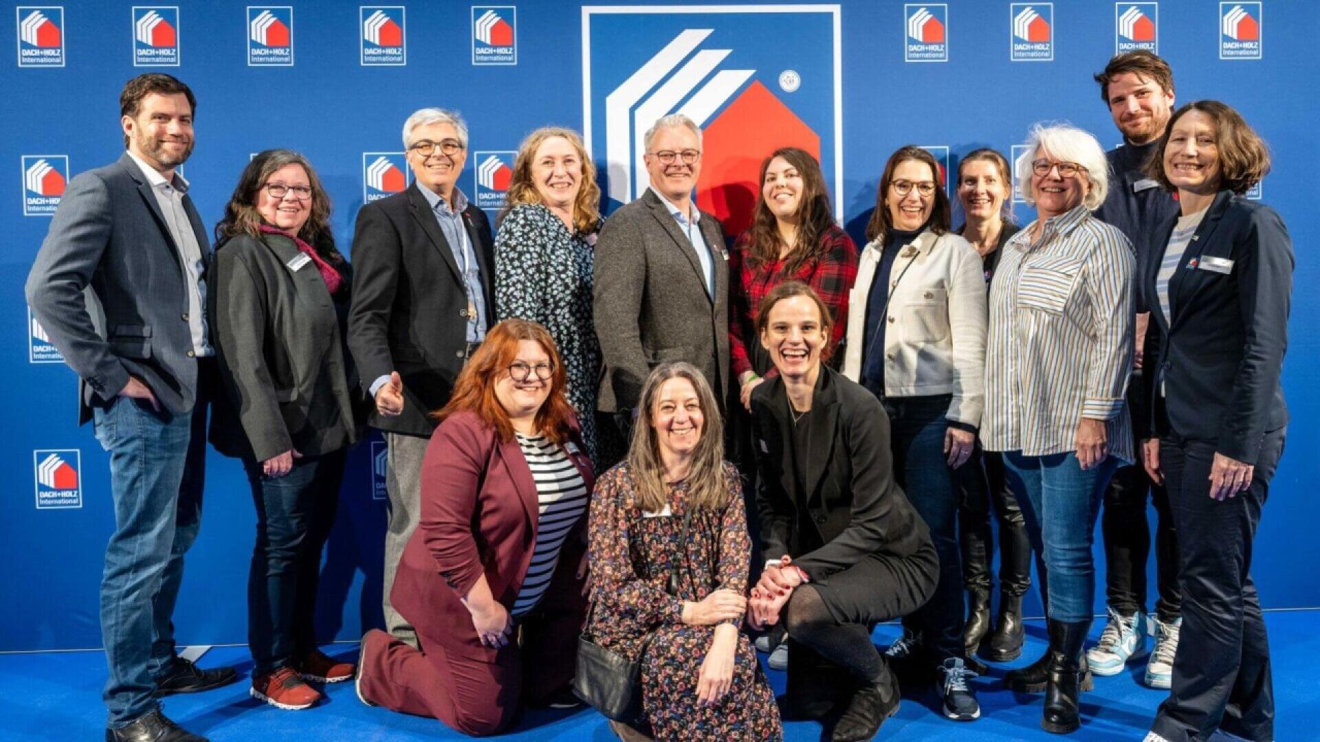 A group of fifteen adults smiling and posing together in front of a blue backdrop with a white and red logo and repeated text. Some are standing, while others kneel in front.