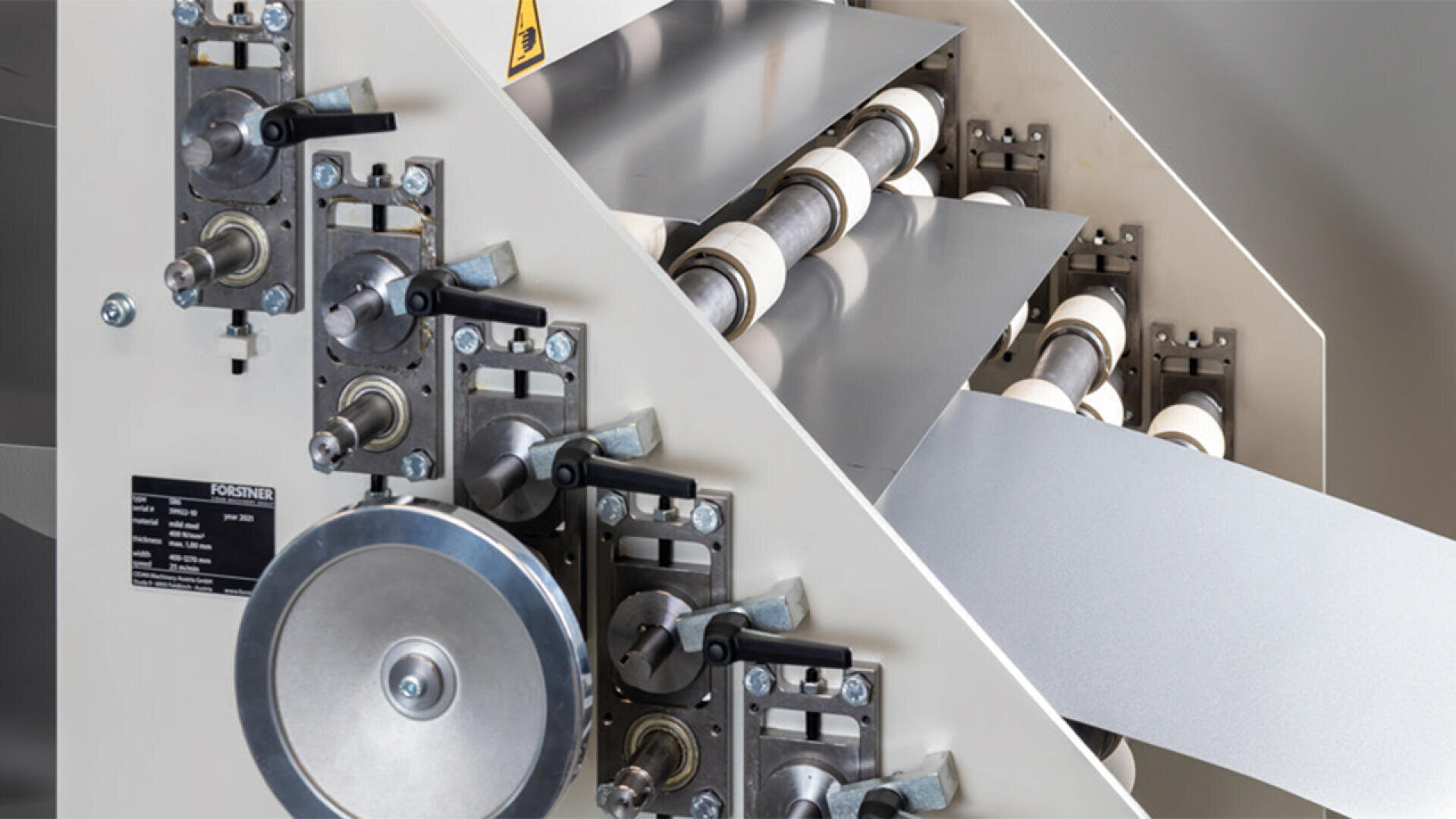Nahaufnahme einer Industriemaschine mit Metallwalzen und Zahnrädern, die ein Materialblatt durch ihren Mechanismus führt. An der Seite des Geräts sind verschiedene Hebel und Einstellregler sichtbar.