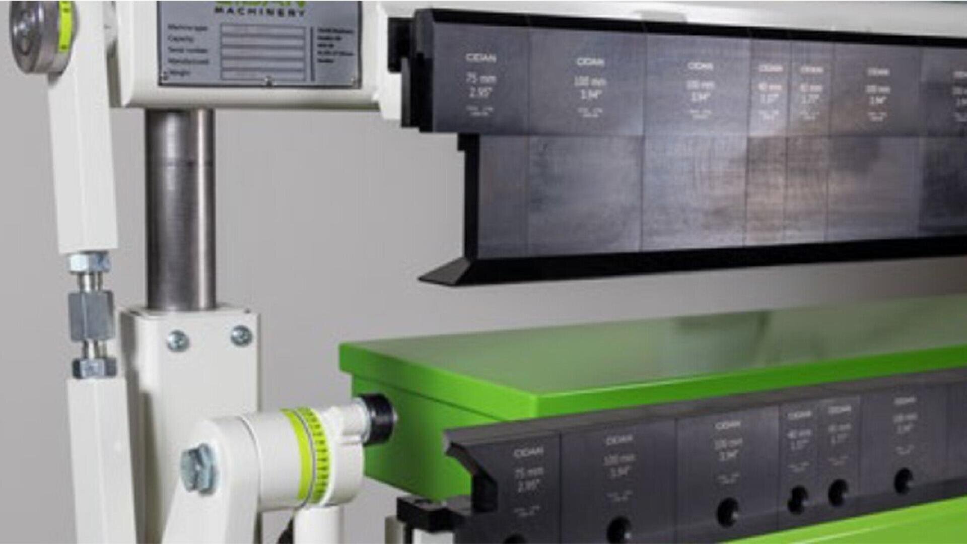 Close-up of industrial sheet metal bending machine with black and green components. Measurement markings are visible on the metal press dies, and a manufacturers label is partially visible on the machine frame.