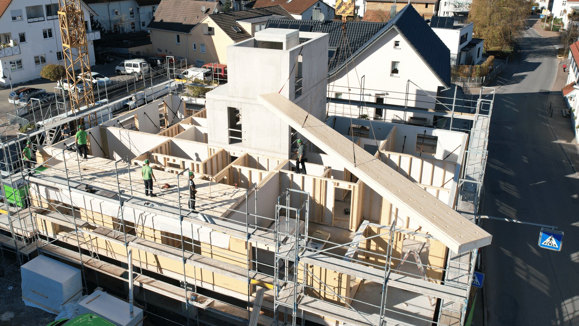 Luftaufnahme eines im Bau befindlichen Gebäudes mit Arbeitern auf der Baustelle, Gerüsten um das Bauwerk herum und Häusern und Straßen in der Nähe. Im oberen Stockwerk wird ein großer Holzbalken angebracht.