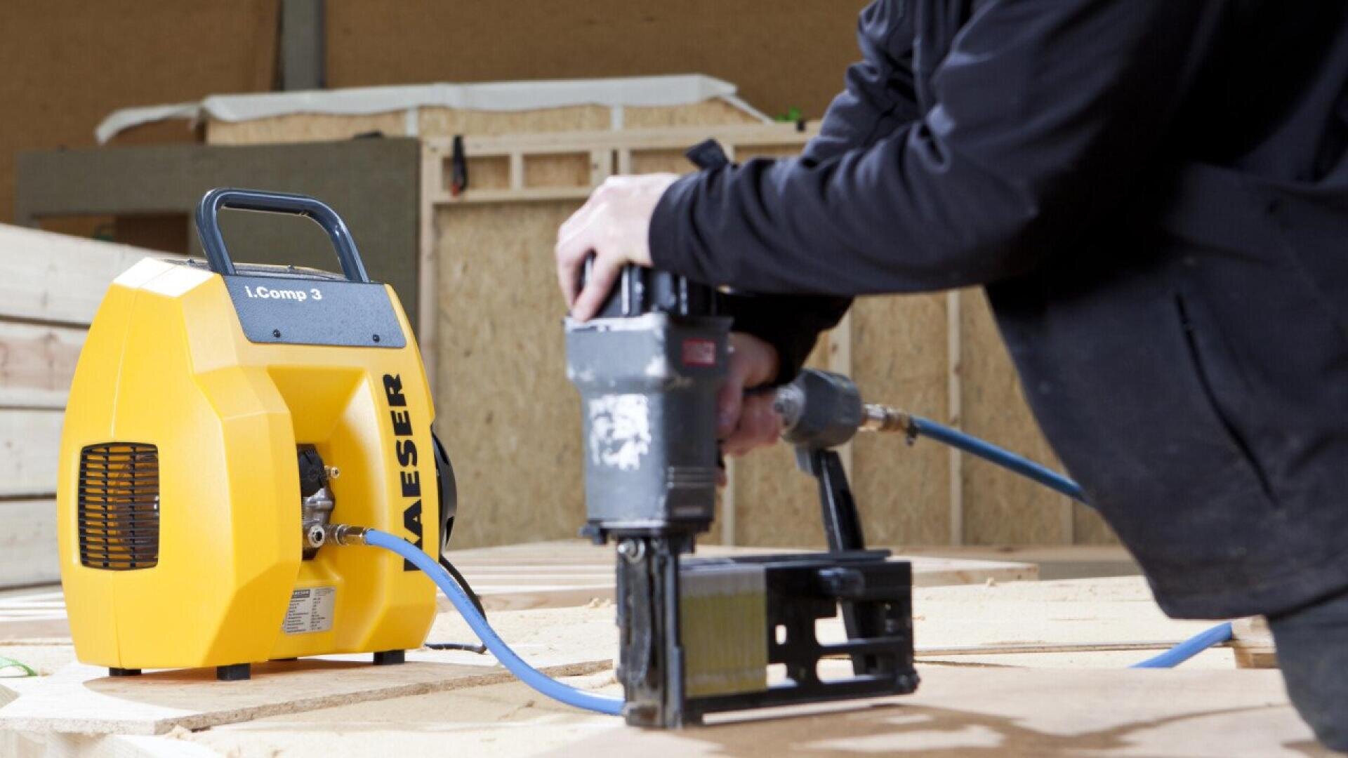 A person uses a pneumatic nail gun connected to a yellow air compressor labeled KAESER while working with wooden boards in a workshop.