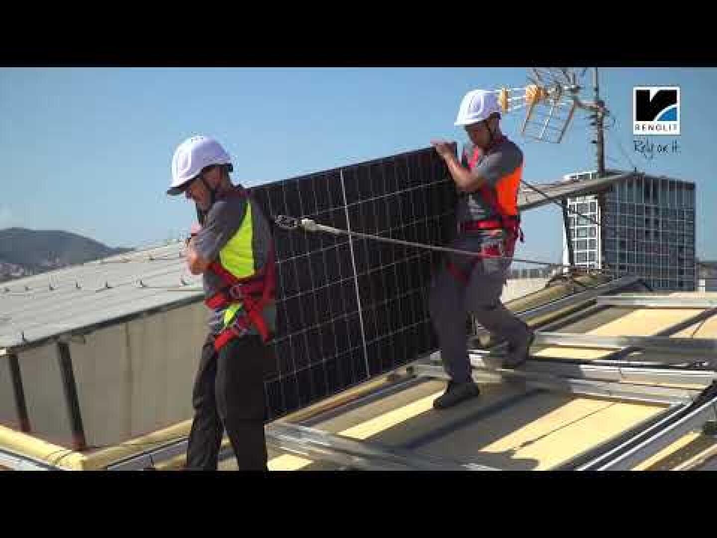Zwei Arbeiter mit Sicherheitsausrüstung und Klettergurten installieren Solarmodule auf einem Dach. Ein Arbeiter hält ein Paneel, während der andere es an seinen Platz führt. Im Hintergrund sind Gebäude und Hügel zu sehen.