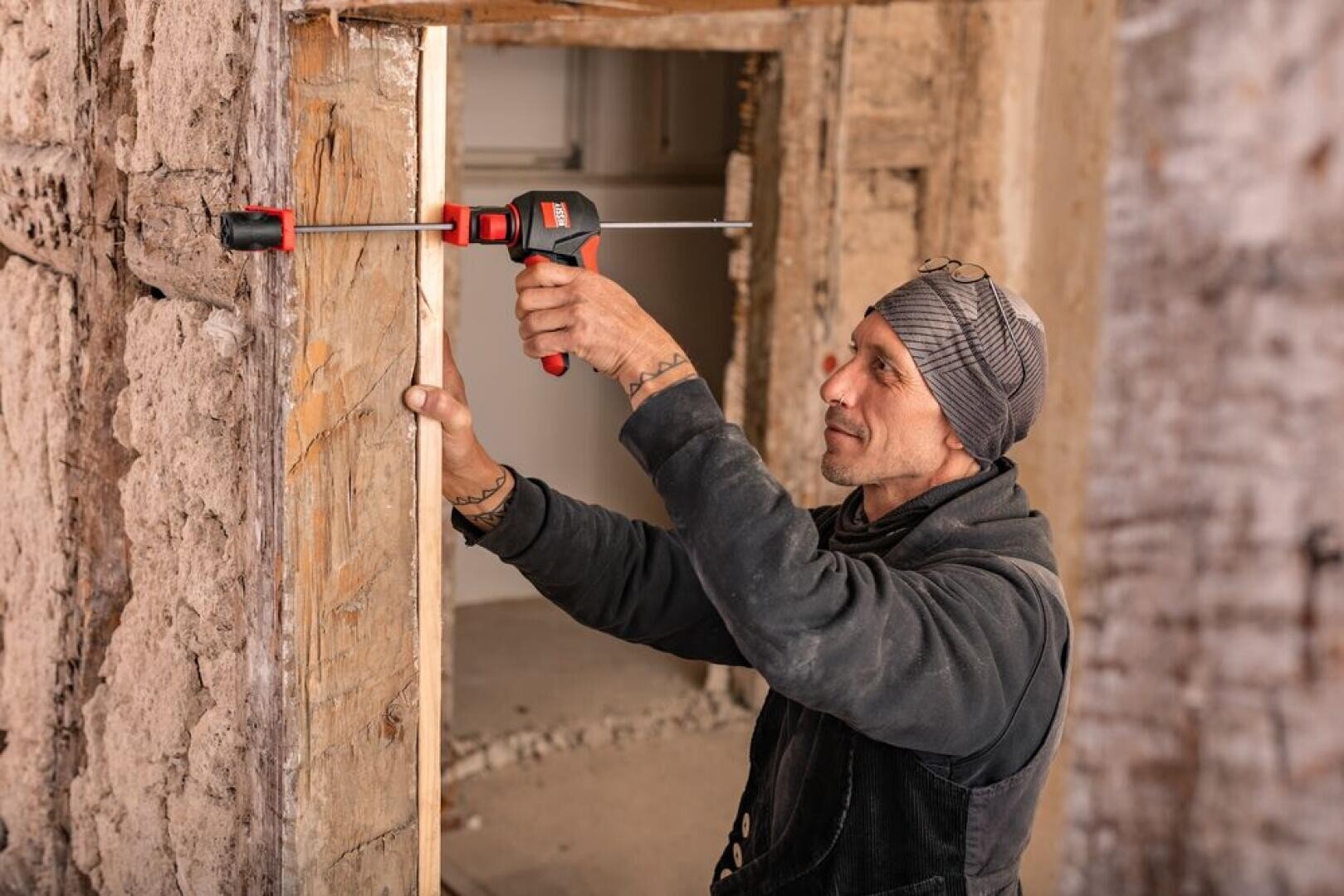 Ein Bauarbeiter mit Mütze benutzt eine Klemme, um ein Holzbrett an einem rauen Türrahmen in einem zu renovierenden Gebäude zu befestigen.