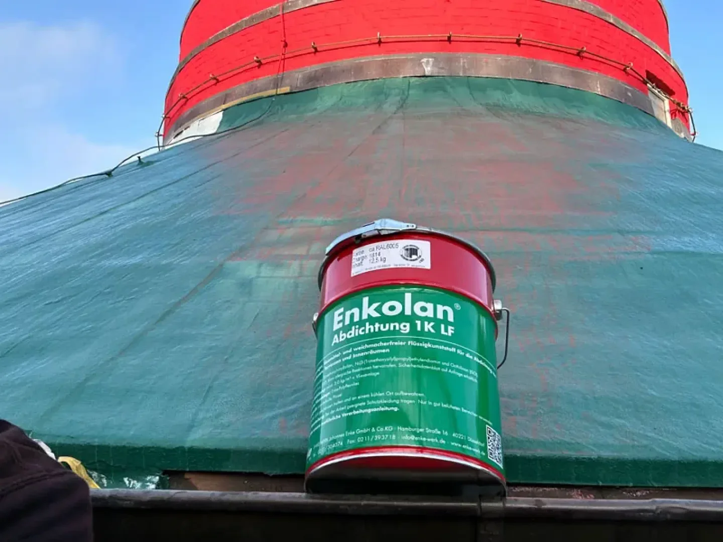 Eine große rot-grüne Dose mit Enkolan-Dichtungsmittel steht auf einem Gerüst vor einer hohen, runden Konstruktion, die mit grüner Plane und rotem Schutzmaterial bedeckt ist.