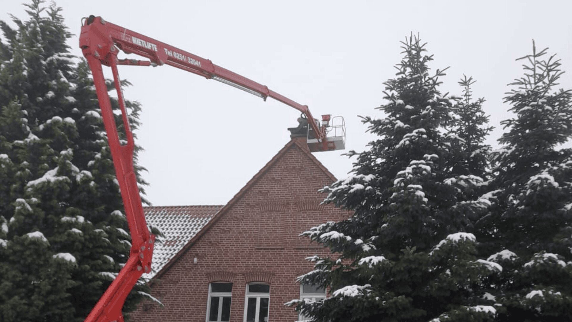 An einem bewölkten Wintertag arbeitet eine Person in einem Korblift in der Nähe des Dachs eines Backsteinhauses, umgeben von hohen, schneebedeckten immergrünen Bäumen.