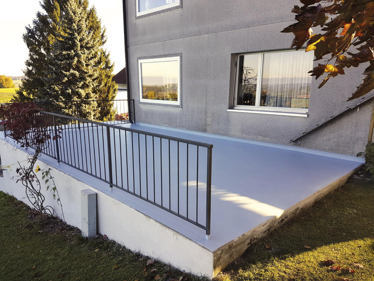 Ein modernes Haus mit grauem Äußeren verfügt über eine erhöhte, flache Terrasse mit glatter Oberfläche und einem schwarzen Metallgeländer, umgeben von grünem Gras und Bäumen.