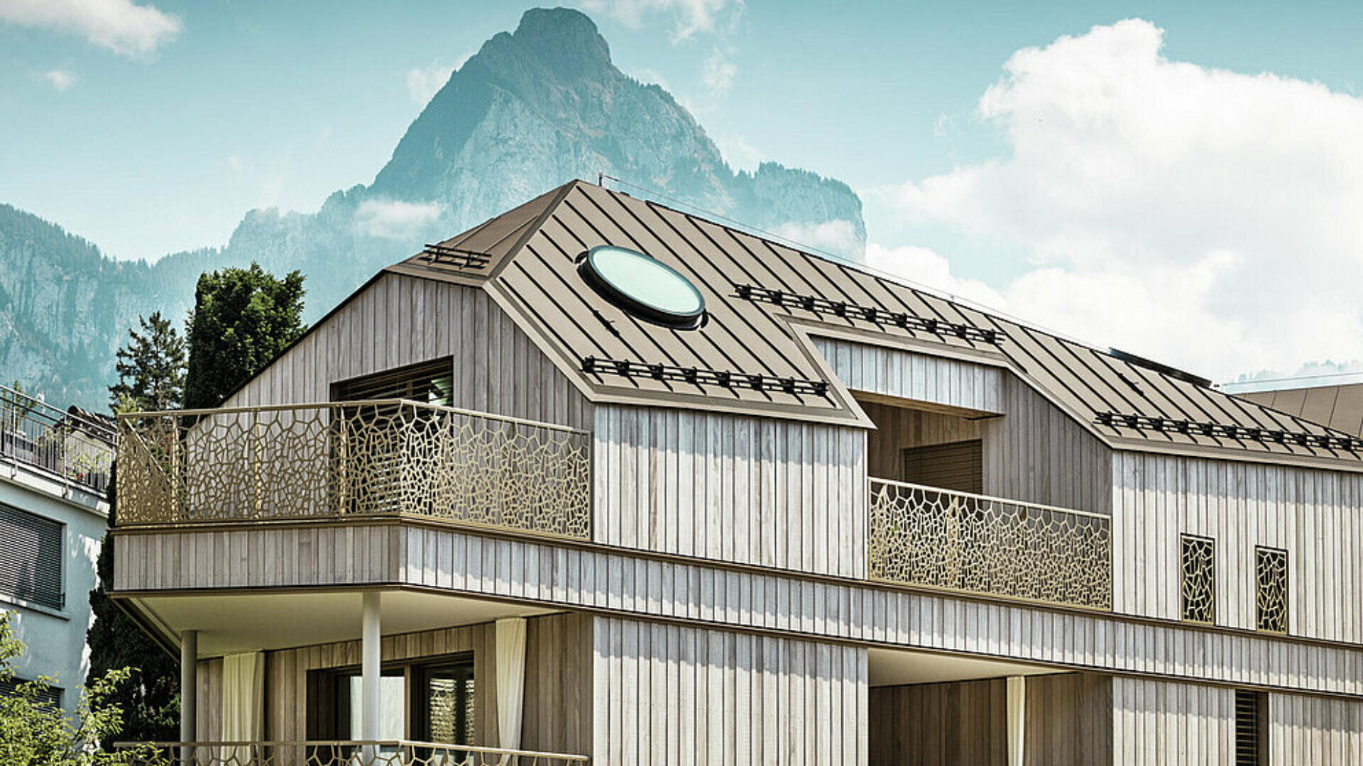 Modernes Holzhaus mit dekorativem Balkongeländer aus Metall, großen Fenstern und einem runden Oberlicht auf dem Schrägdach, vor der Kulisse hoher Berge und teilweise bewölktem Himmel.