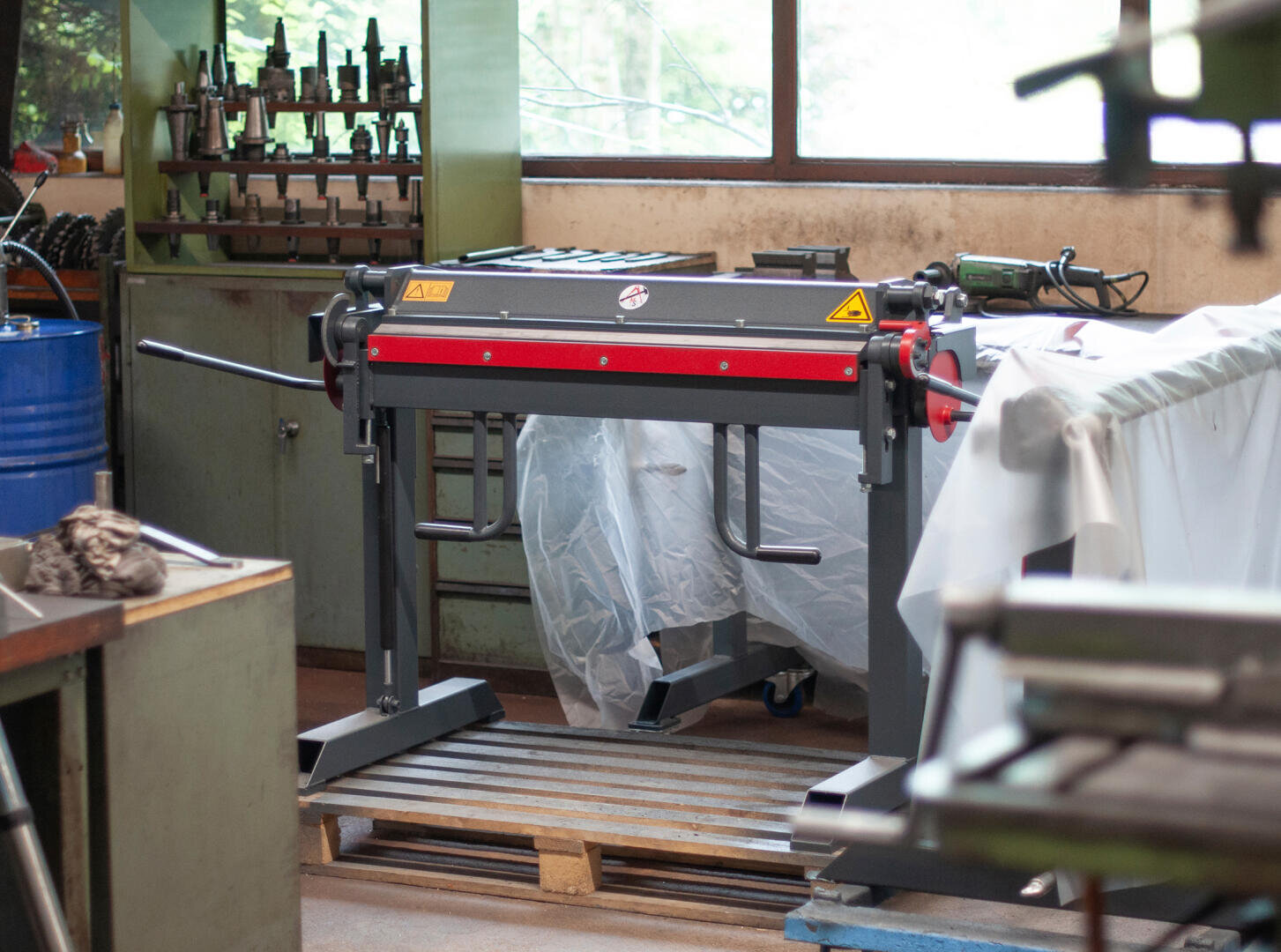 A metal bending machine stands in a workshop surrounded by tools, machines and covered equipment, with natural light streaming in through the large windows.