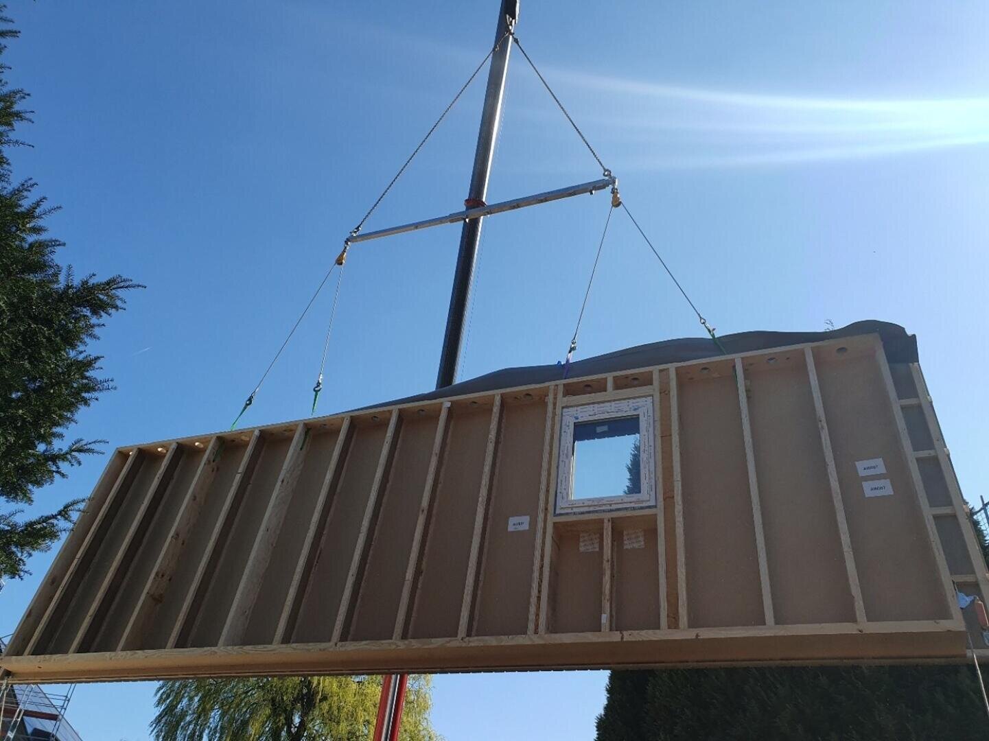 Ein vorgefertigtes hölzernes Wandelement mit einer Fensteröffnung wird von einem Kran vor einem strahlend blauen Himmel angehoben, wahrscheinlich zum Einbau auf einer Baustelle.