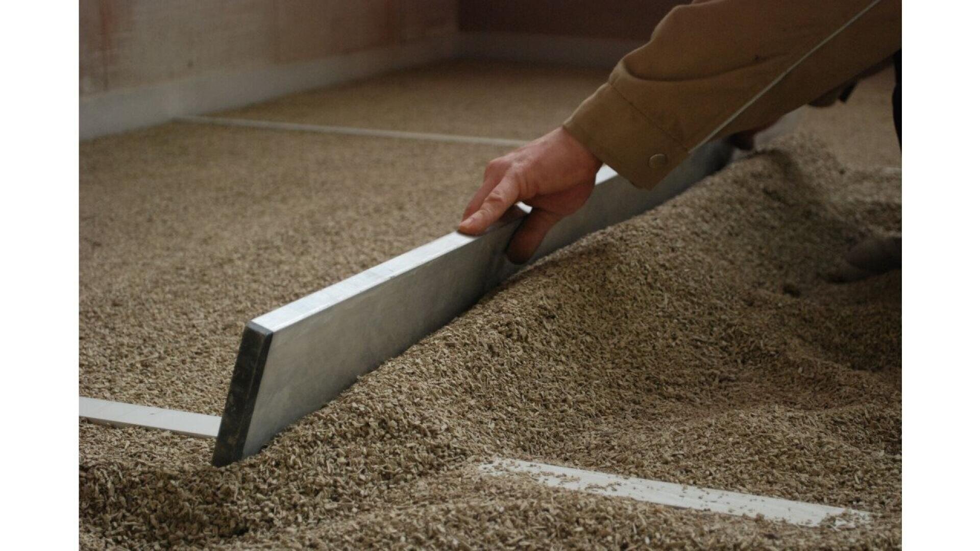 A person uses a straight metal tool to level a layer of loose gravel or aggregate material on the ground, preparing an even surface.