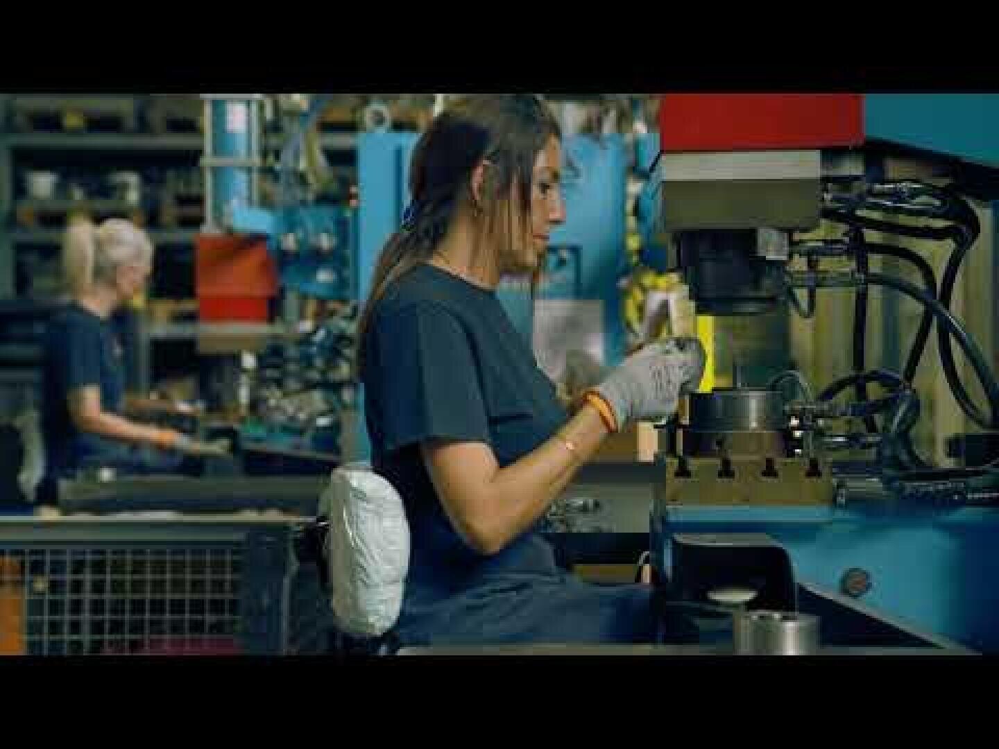 Eine Person mit Handschuhen bedient eine Maschine in einer Fabrik, während im Hintergrund Regale und ein weiterer Arbeiter zu sehen sind.