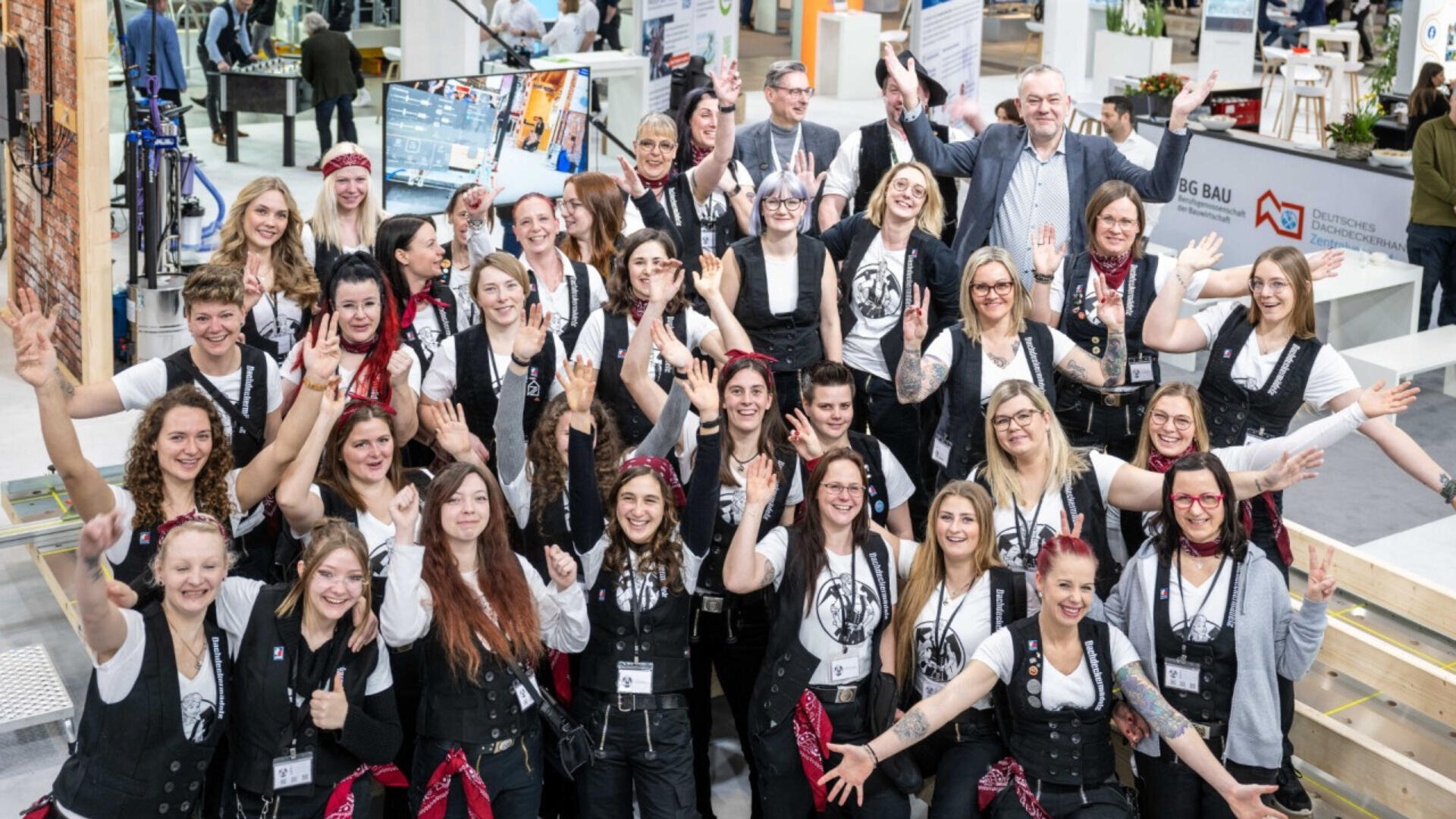 Eine große Gruppe fröhlicher Menschen, hauptsächlich Frauen in passenden Westen und weißen Hemden, posiert gemeinsam bei einer Veranstaltung in geschlossenen Räumen. Viele von ihnen lächeln und heben aufgeregt die Arme.