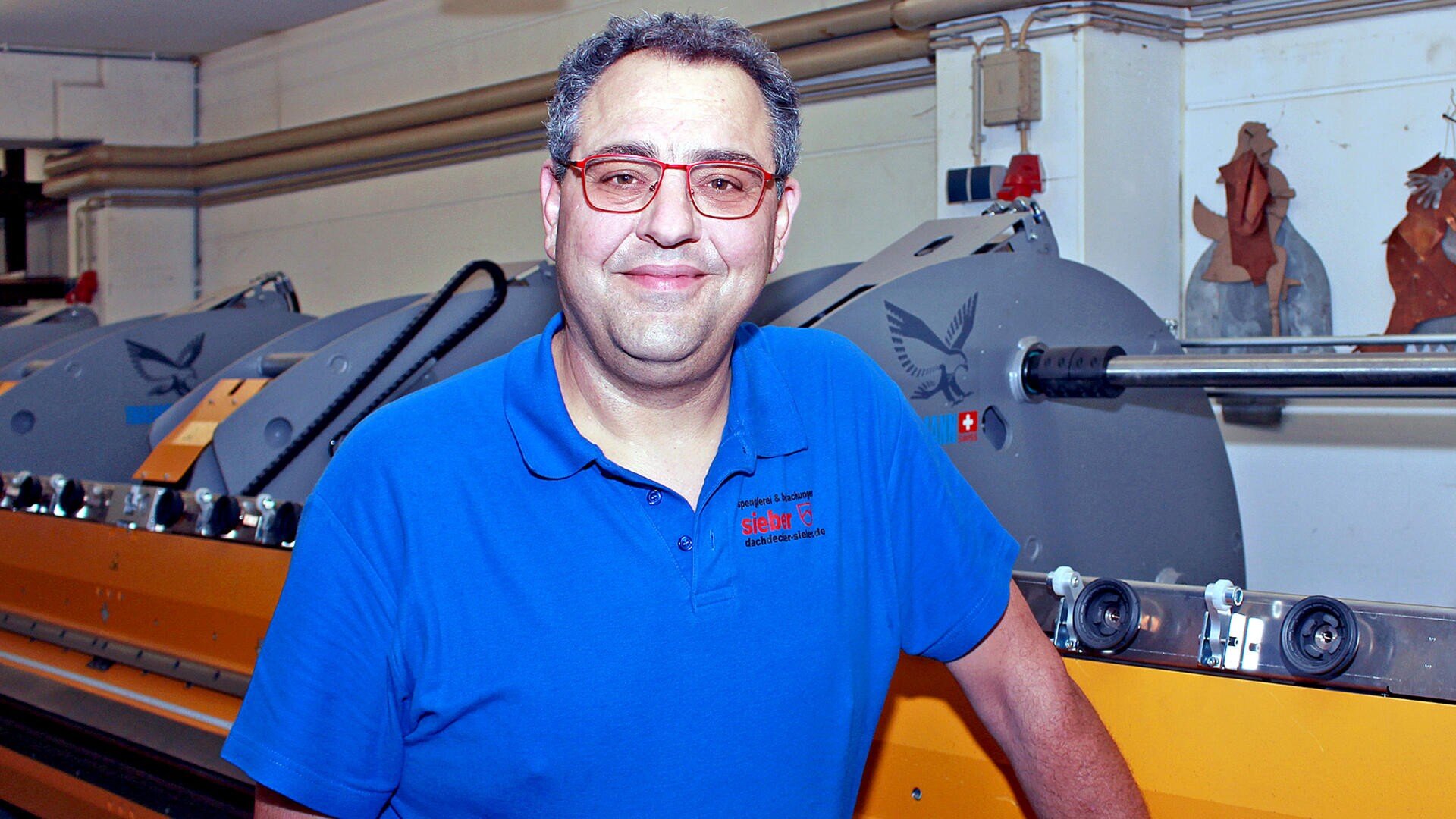 Ein lächelnder Mann mit Brille und blauem Poloshirt steht in einem hell erleuchteten Innenraum vor großen Industriemaschinen.