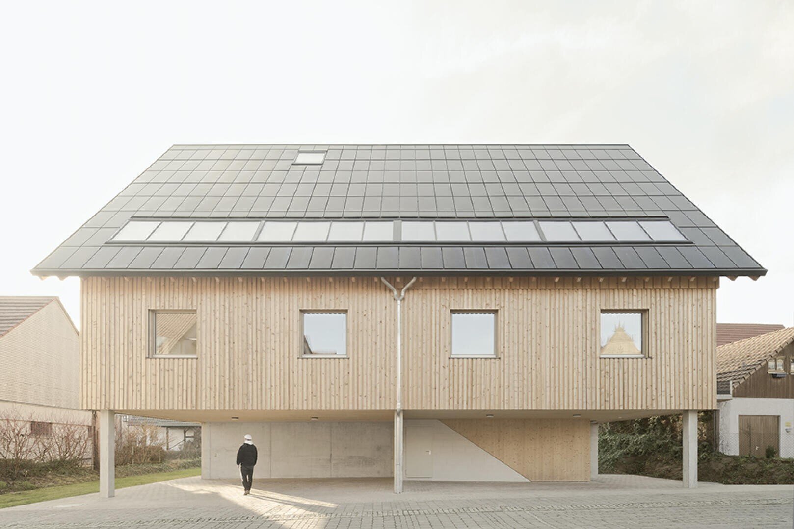 A modern wooden house on stilts with a large solar cell roof. A person walks under the elevated building to the entrance. The house has several windows and an open parking lot underneath.
