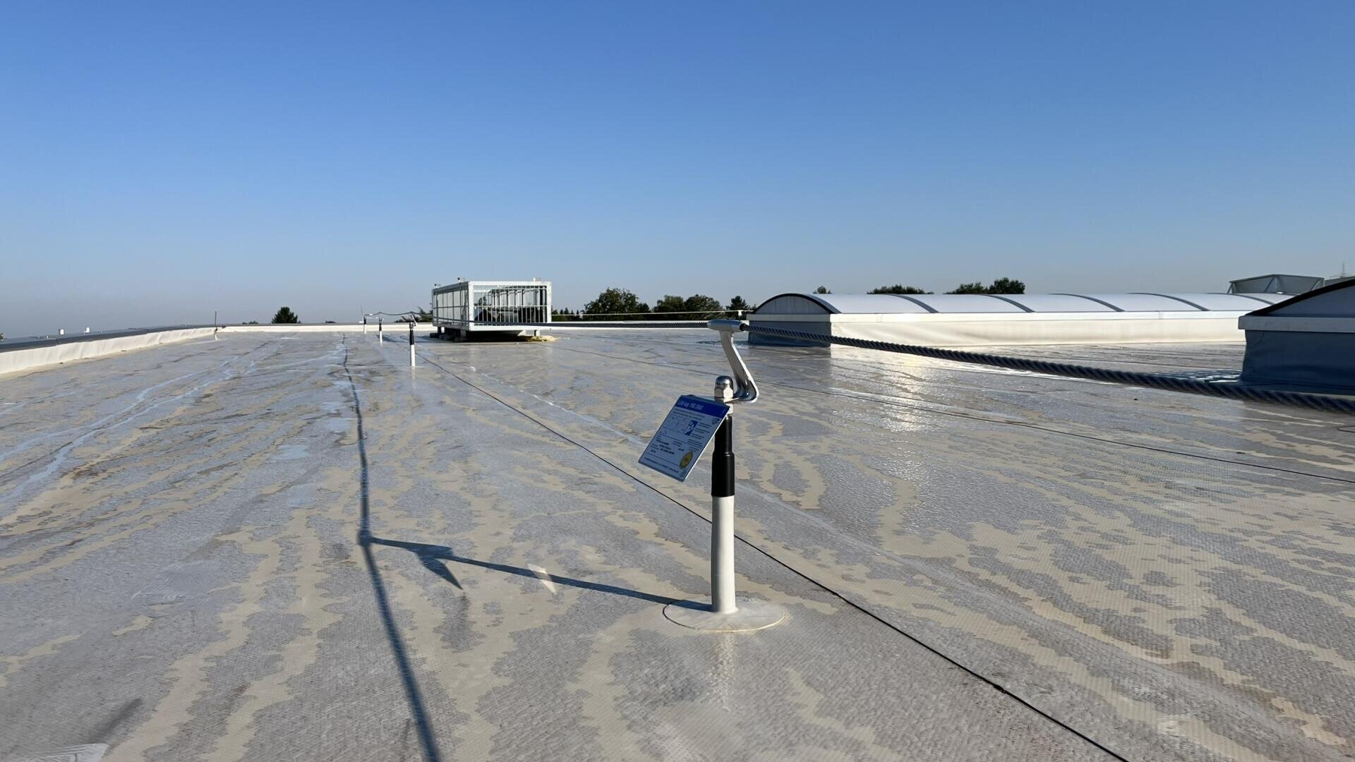 Ein flaches, sonnenbeschienenes Dach mit hellen Abdichtungen, Sicherheitsleinenpfosten und einem Warnschild im Vordergrund. Im Hintergrund sind Oberlichter, Lüftungsöffnungen und ein klarer blauer Himmel zu sehen.