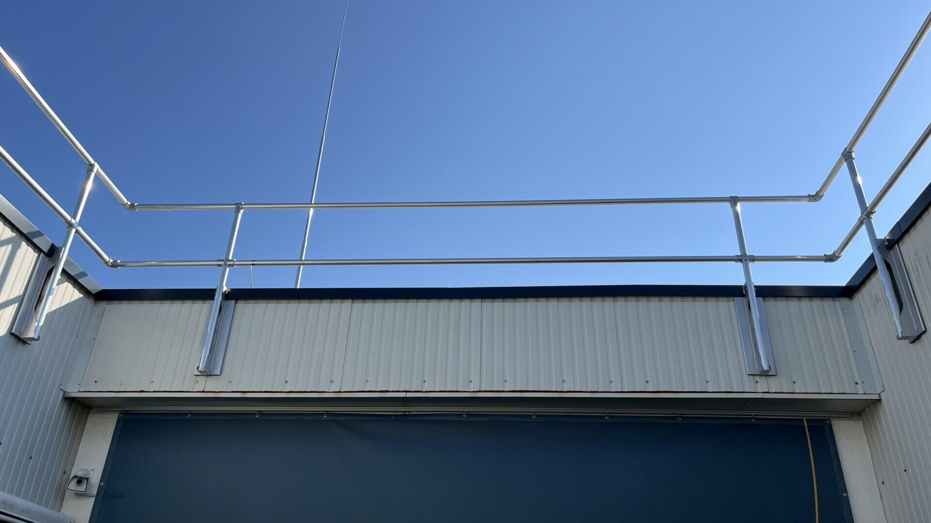 Ein klarer blauer Himmel über einem flachen Dach mit einem Sicherheitsgeländer aus Metall. Das Gebäude hat helle Seitenwände und einen dunkelblauen Bereich unterhalb des Geländers. Im Hintergrund ragt eine dünne Antenne in die Höhe.