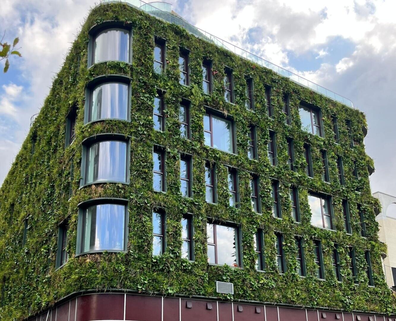 Ein modernes Gebäude mit großen Fenstern und mit Grünpflanzen bewachsenen Wänden, die einen vertikalen Garteneffekt erzeugen; im Hintergrund ist der Himmel mit einigen Wolken zu sehen.