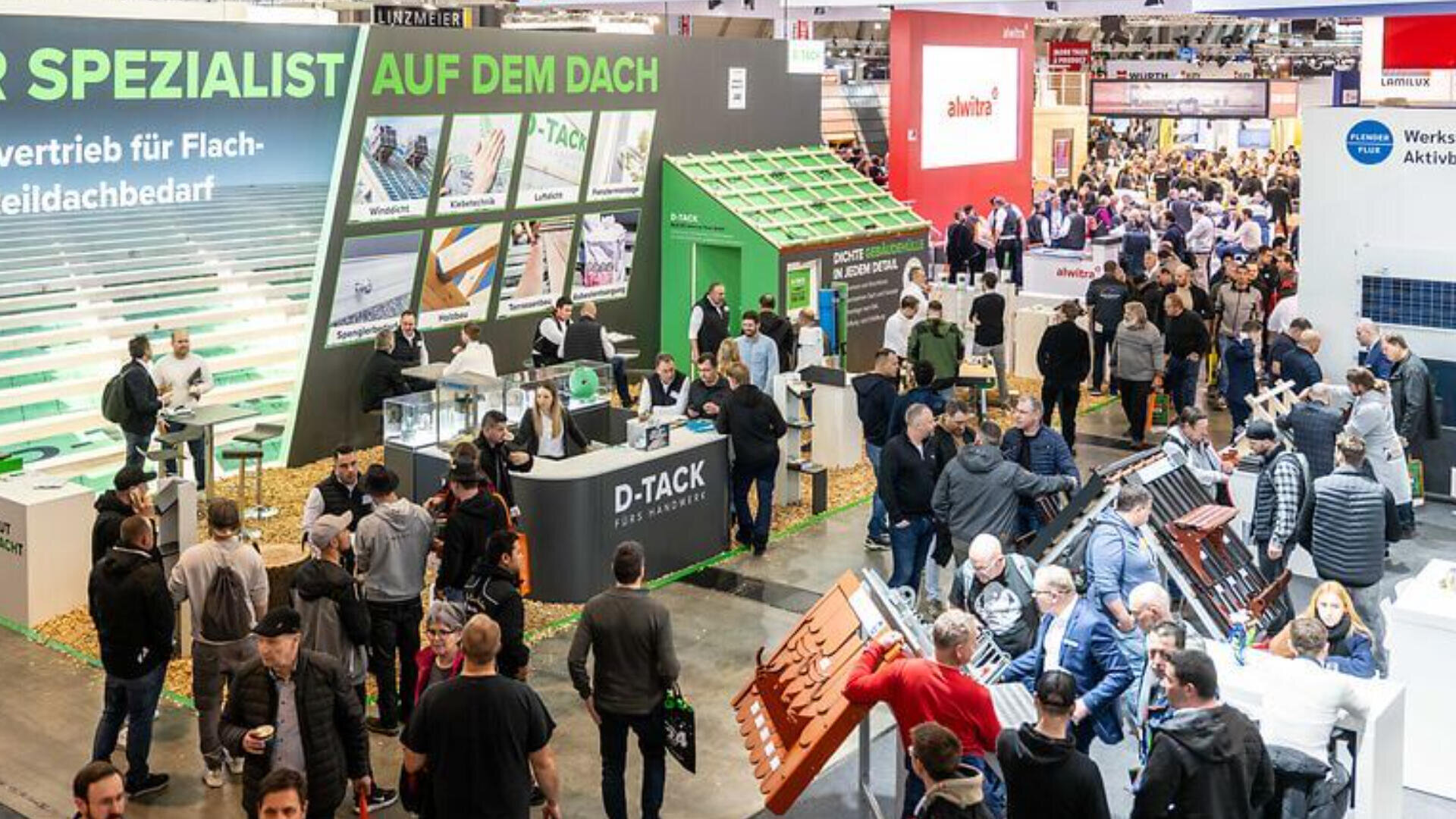 Eine gut besuchte Messe mit vielen Besuchern, die Stände besuchen und sich mit den Ausstellungen zu Dach- und Baumaterialien beschäftigen. In der gesamten Messehalle sind verschiedene Firmenschilder, Demo-Aufbauten und Produktmuster zu sehen.