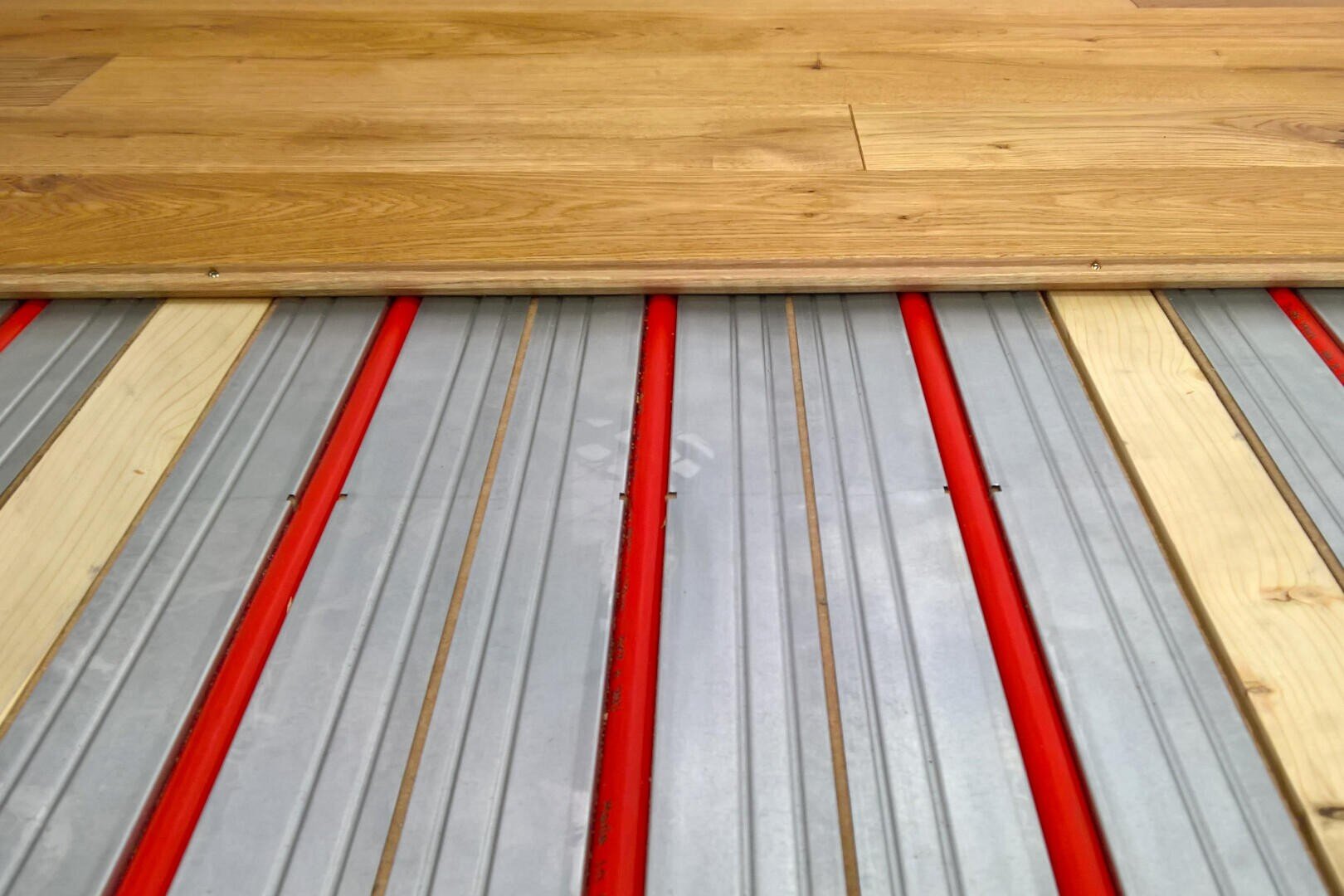 Nahaufnahme eines teilweise verlegten Holzbodens mit roten Fußbodenheizungsrohren in Metallkanälen unter den Dielen.