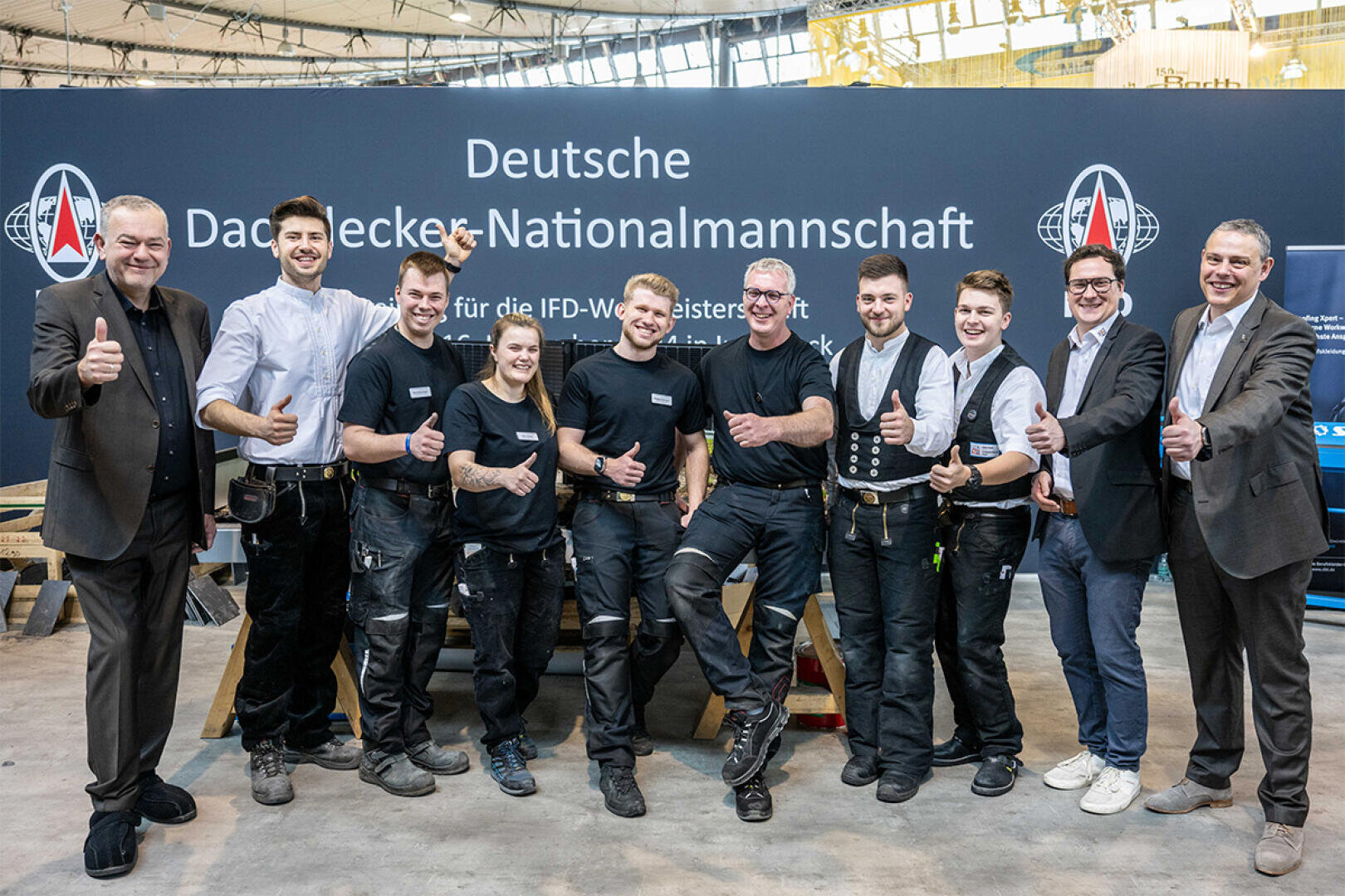 Eine Gruppe von zehn Personen, einige in Arbeitsuniformen, andere in Geschäftskleidung, steht lächelnd und mit erhobenem Daumen vor einem Schild mit der Aufschrift Deutsche Dachdecker Nationalmannschaft.